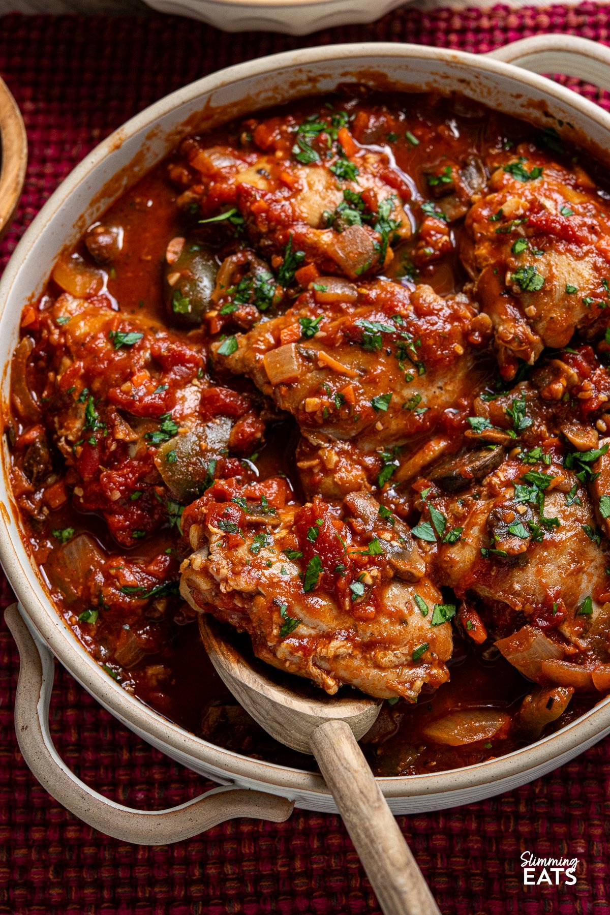 Transform your family meals with Slow Cooker Chicken Cacciatore. Tender chicken thighs meld beautifully with green bell pepper and mushrooms in a rich tomato sauce. Serve it with your favourite sides for a great meal that everyone will enjoy. close up of chicken cacciatore in double handled casserole dish with wooden spoon