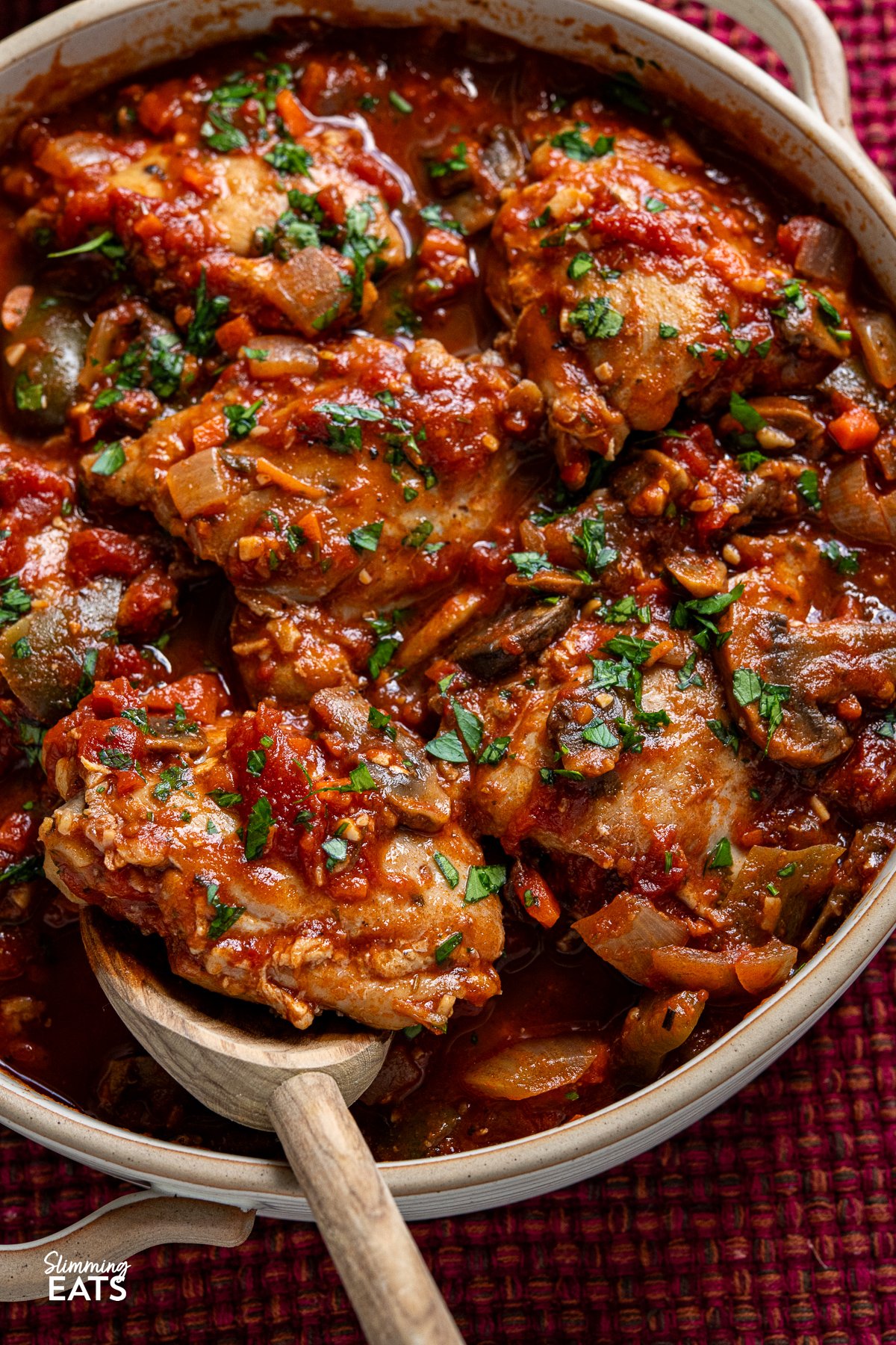 Transform your family meals with Slow Cooker Chicken Cacciatore. Tender chicken thighs meld beautifully with green bell pepper and mushrooms in a rich tomato sauce. Serve it with your favourite sides for a great meal that everyone will enjoy. close up of chicken cacciatore in double handled casserole dish with wooden spoon