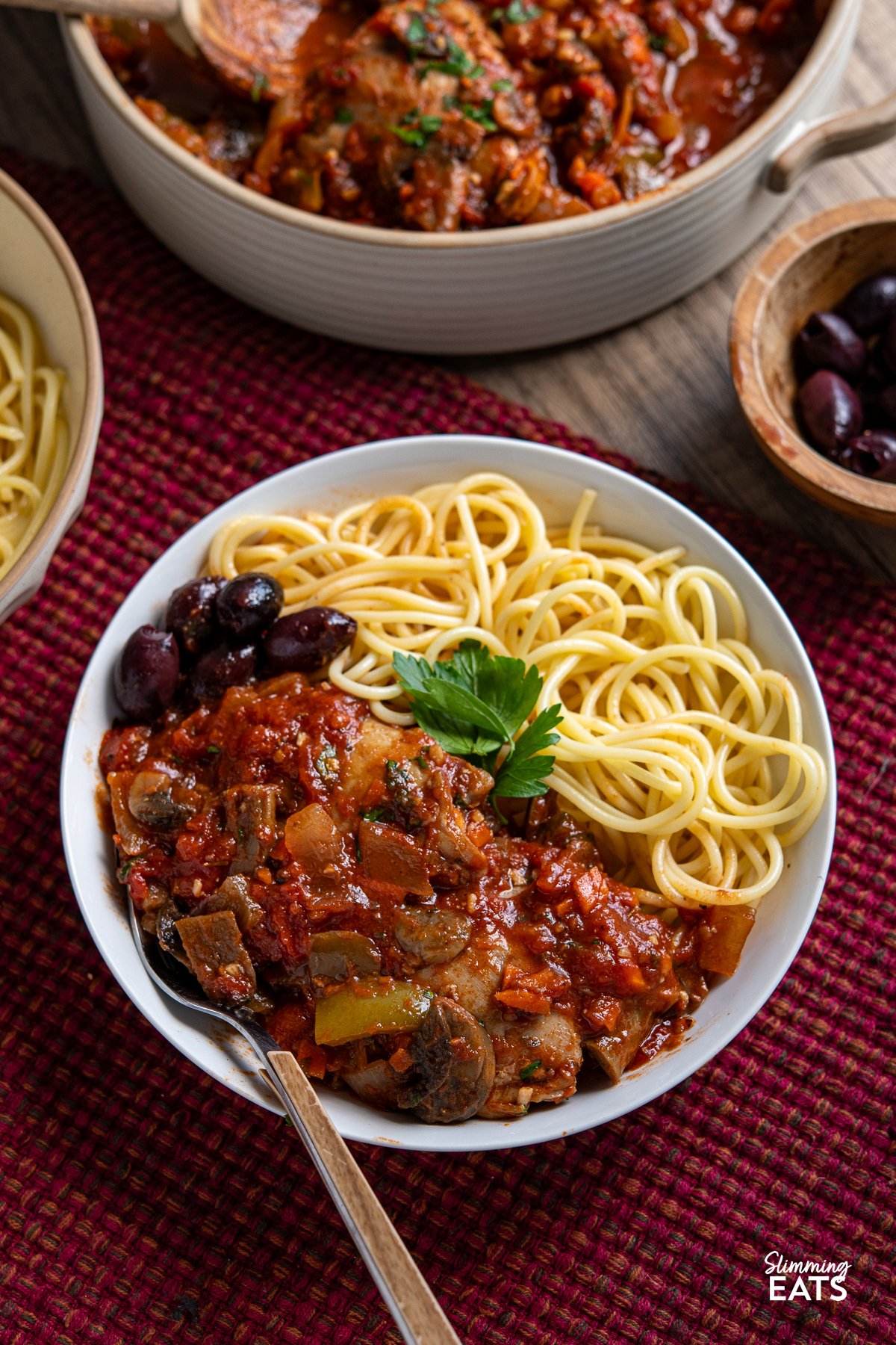 Transform your family meals with Slow Cooker Chicken Cacciatore. Tender chicken thighs meld beautifully with green bell pepper and mushrooms in a rich tomato sauce. Serve it with your favourite sides for a great meal that everyone will enjoy. chicken cacciatore in white bowl with spaghetti and black olives, fork placed in left hand side of bowl
