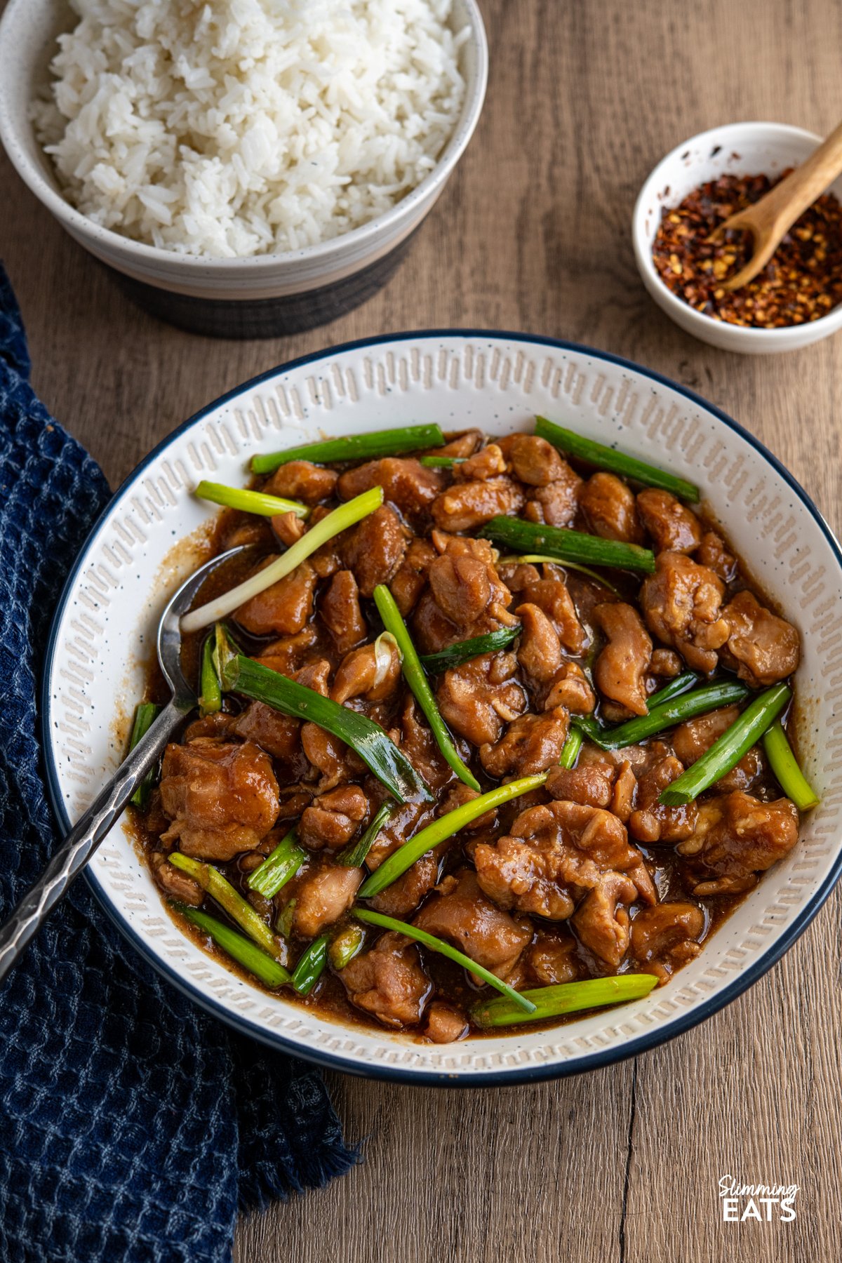 Slow Cooker Mongolian Chicken is a fuss-free dinner that’s brimming with bold, savoury-sweet flavours. Tender chicken is coated in a rich, glossy sauce made with soy, garlic, and a touch of brown sugar, creating a melt-in-your-mouth meal the whole family will love. Pair it with fluffy jasmine rice for a comforting dish that tastes like a takeaway classic! slow cooker mongolian chicken in a white bowl with navy blue rim, bowl of jasmine rice and small bowl of chilli flakes in background