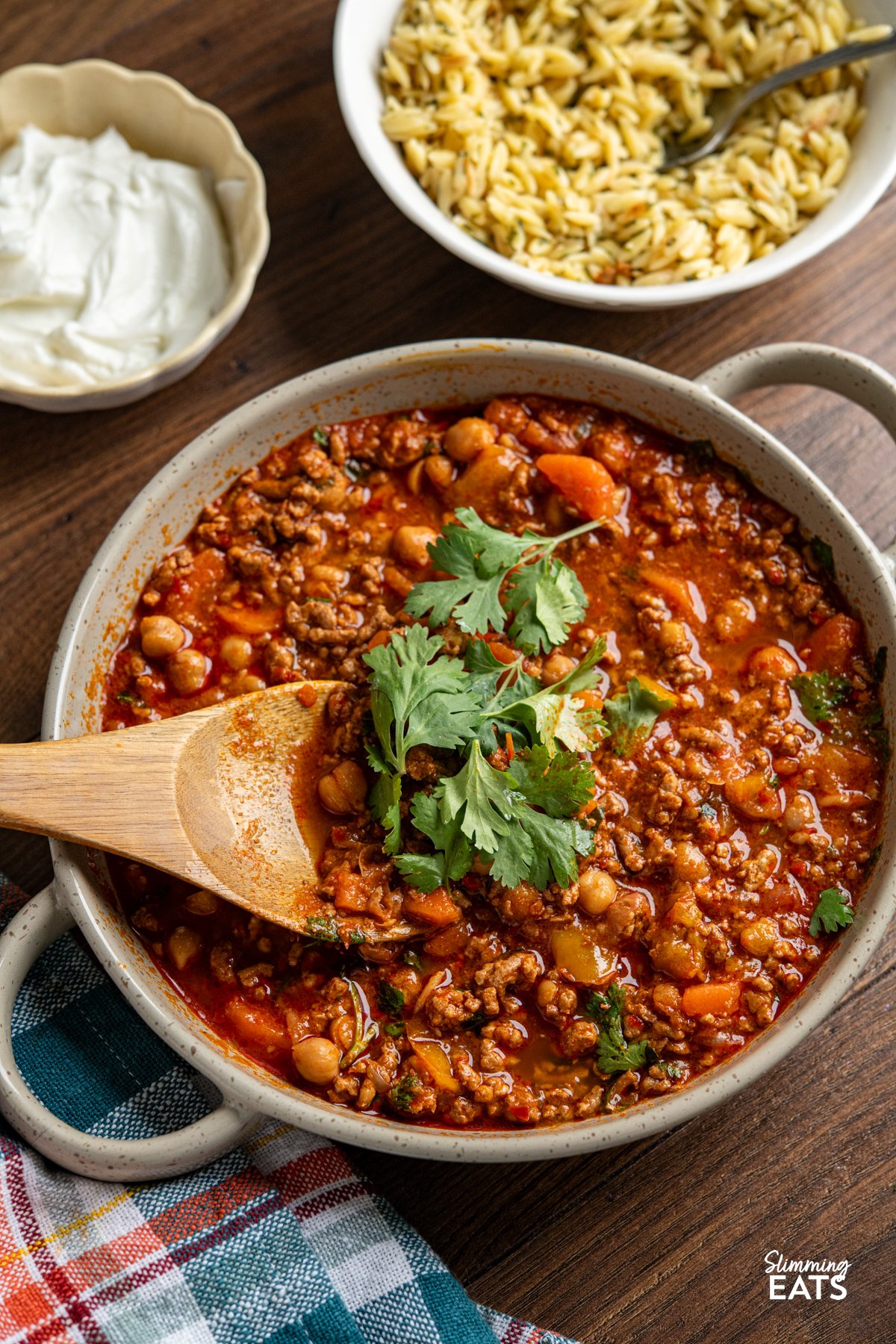 Let your slow cooker do the work with this flavour-packed Harissa Ground Pork, where savoury pork simmers with chickpeas, sweet carrots, and peppers in a rich, spiced sauce. A touch of harissa brings warmth without overpowering, while garlic, cumin, and smoked and sweet paprika add depth to every bite. This effortless dish comes together beautifully, making it a perfect choice for a hearty, satisfying meal. Slow Cooker Harissa Ground Pork in a round grey brown double handled ceramic dish with wooden spoon - fresh coriander added as a garnish. Orzo and Greek yoghurt surrounds the dish in bowls.