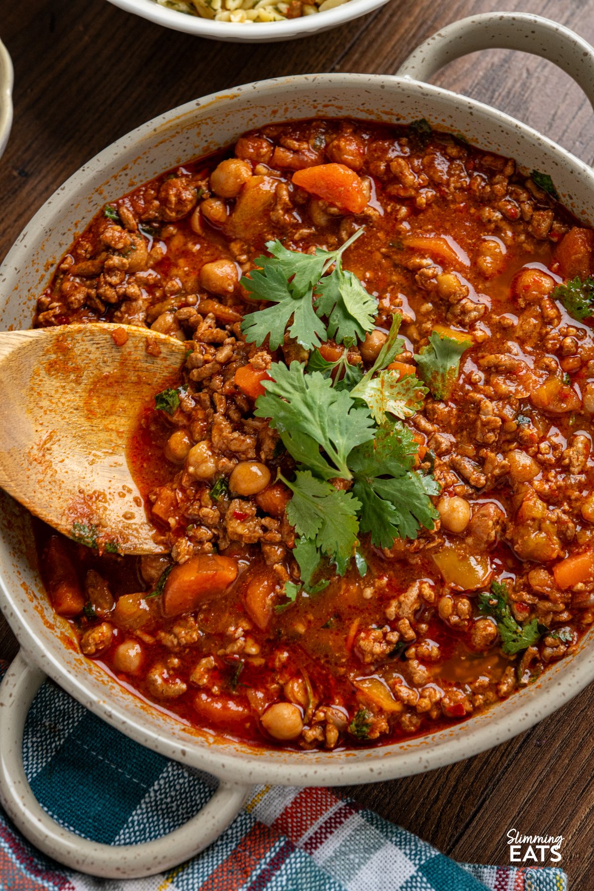 Let your slow cooker do the work with this flavour-packed Harissa Ground Pork, where savoury pork simmers with chickpeas, sweet carrots, and peppers in a rich, spiced sauce. A touch of harissa brings warmth without overpowering, while garlic, cumin, and smoked and sweet paprika add depth to every bite. This effortless dish comes together beautifully, making it a perfect choice for a hearty, satisfying meal. close up of Slow Cooker Harissa Ground Pork in a round grey brown double handled ceramic dish with wooden spoon - fresh coriander added as a garnish.