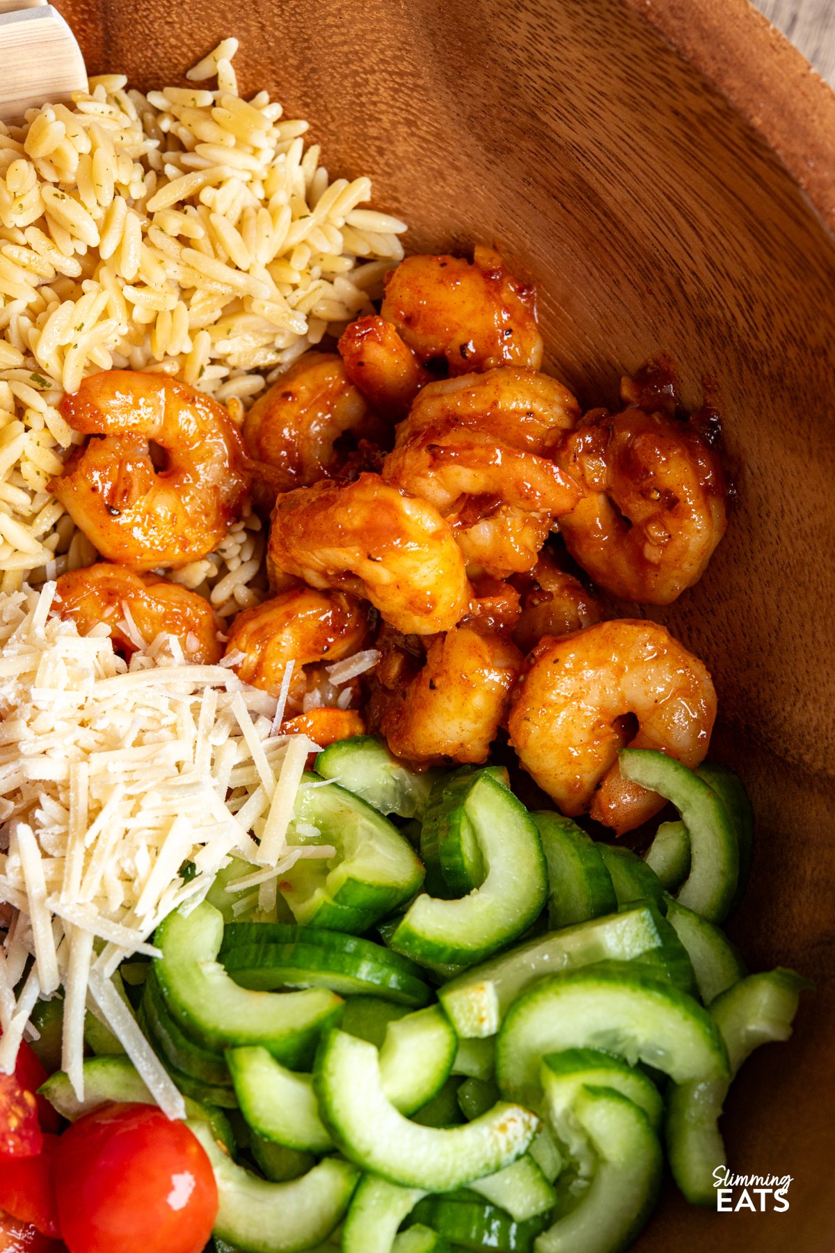 Fresh, vibrant, and packed with flavour—this Shrimp Orzo Salad is a summer stunner. Tender orzo tossed with crisp cucumber, juicy cherry tomatoes, and slivers of red onion forms the perfect base for buttery prawns, pan-fried with garlic, parsley, paprika and a hint of chilli. A drizzle of honey, a squeeze of lime, and a generous grating of Parmesan tie it all together with a sweet-savoury kick. close up of Easy Shrimp Orzo Salad in a wooden salad bowl ready to be mixed