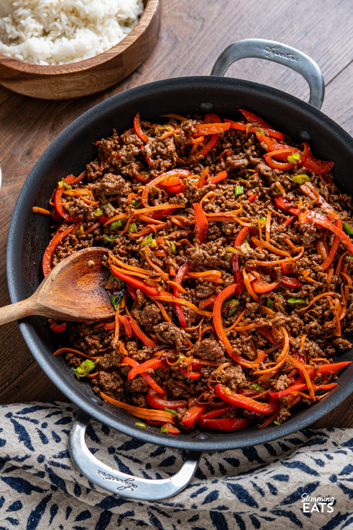 Sweet, savoury, and just the right amount of heat—this Spicy Sticky Ground Beef is a flavour-packed, fuss-free dish. Minced beef is browned and simmered in a glossy sauce of soy, sriracha, maple syrup, and sweet chilli, with garlic and ginger adding warmth. Red bell pepper and carrot bring a hint of sweetness and a satisfying bite, making every mouthful rich, sticky, and completely irresistible. Spicy Sticky Ground Beef in a double handled large non-stick skillet, rice in wooden bowl visible
