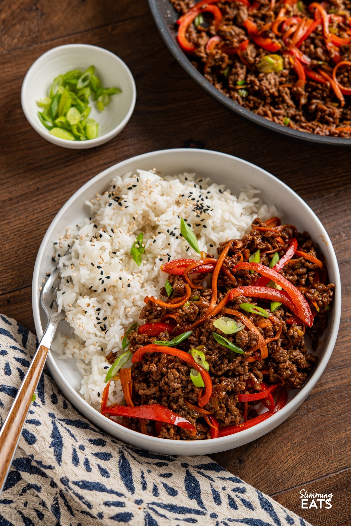 Sweet, savoury, and just the right amount of heat—this Spicy Sticky Ground Beef is a flavour-packed, fuss-free dish. Minced beef is browned and simmered in a glossy sauce of soy, sriracha, maple syrup, and sweet chilli, with garlic and ginger adding warmth. Red bell pepper and carrot bring a hint of sweetness and a satisfying bite, making every mouthful rich, sticky, and completely irresistible. Spicy Sticky Ground Beef in a white bowl with jasmine rice, fork placed in left side of bowl, pan visible in background with small bowl of chopped spring onions