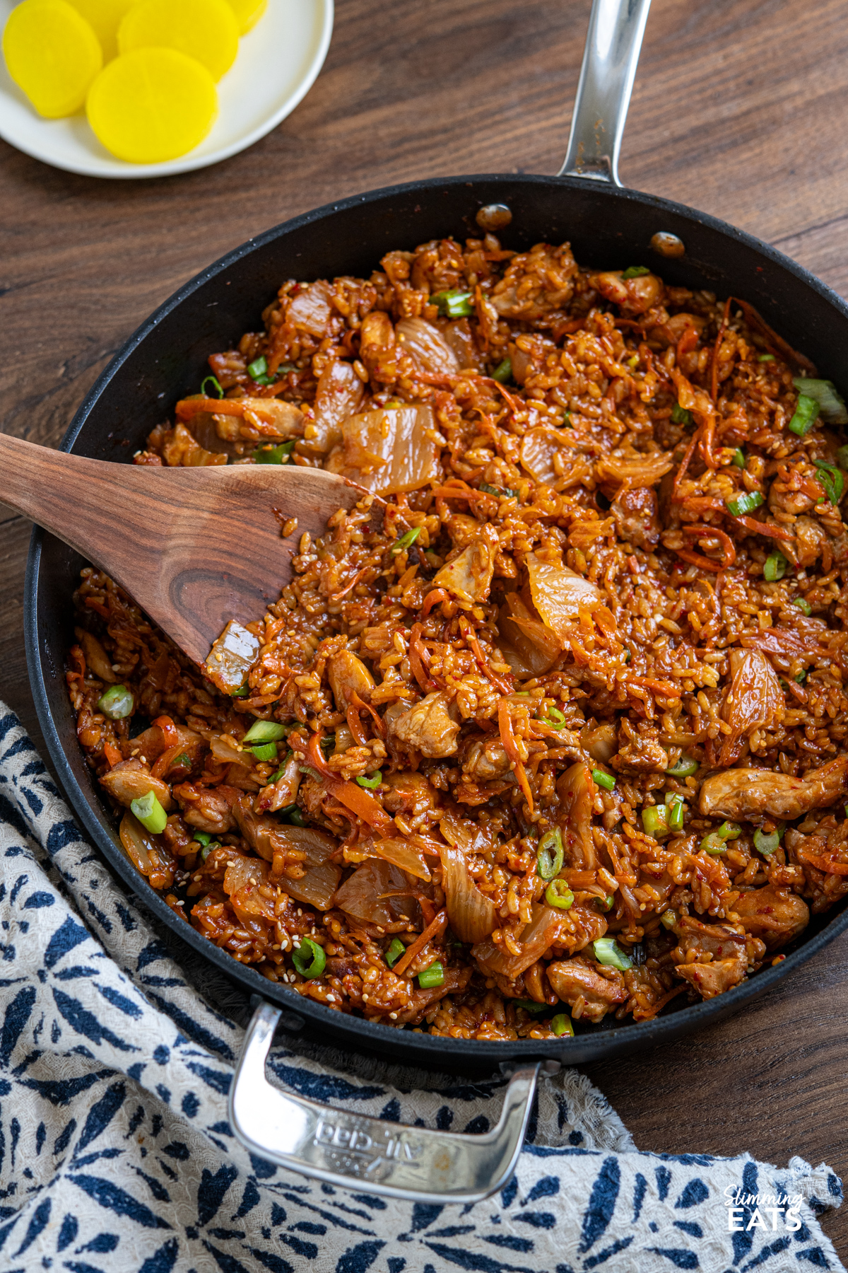 Spicy, savoury, and utterly moreish—this One Pot Chicken Kimchi Rice is a weeknight winner with bold, punchy flavour and minimal fuss. Tender chicken thighs simmer gently with tangy kimchi and a touch of sweetness, creating a rich, satisfying dish that brings comforting Korean-inspired vibes to your dinner table. One Pot Chicken Kimchi Rice in all clad frying pan with small plate of yellow pickled radish in background.