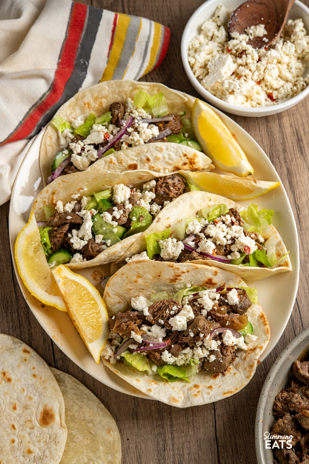 This Slow Cooker Greek Lamb is packed with bold, Mediterranean-inspired flavour and couldn’t be easier to make. Lamb is slow-cooked with onion, garlic, lemon juice and a fragrant mix of rosemary, thyme, oregano and cumin until it’s melt-in-your-mouth tender. A touch of paprika and red pepper flakes adds gentle warmth, while stock keeps everything juicy and full of flavour. slow cooker greek lamb mino soft tortilla tacos on a oval plate topped with lettuce, cucumber, red onion and crumbled feta, lemon wedges on the side.