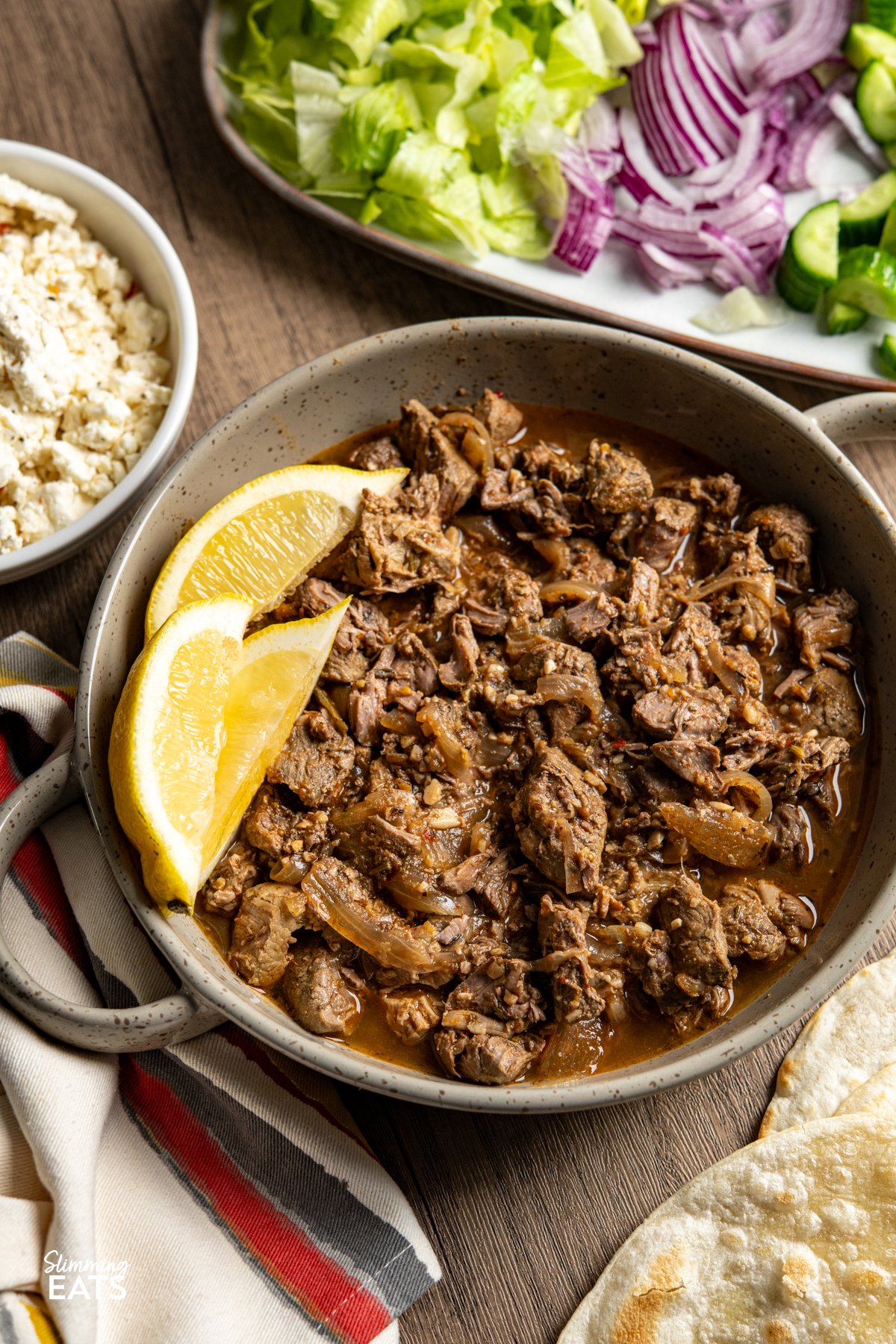 This Slow Cooker Greek Lamb is packed with bold, Mediterranean-inspired flavour and couldn’t be easier to make. Lamb is slow-cooked with onion, garlic, lemon juice and a fragrant mix of rosemary, thyme, oregano and cumin until it’s melt-in-your-mouth tender. A touch of paprika and red pepper flakes adds gentle warmth, while stock keeps everything juicy and full of flavour. slow cooker greek lamb in double handled beige brown ceramic dish with lemon wedges surrounded by feta and salad.