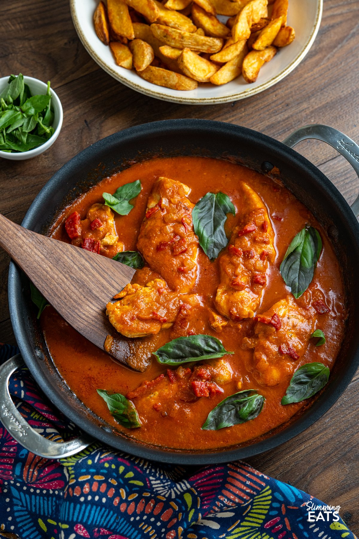 Chicken in Roasted Red Pepper Basil Sauce in double handled non stick pan with a bowl of potato wedges and small bowl of fresh basil 