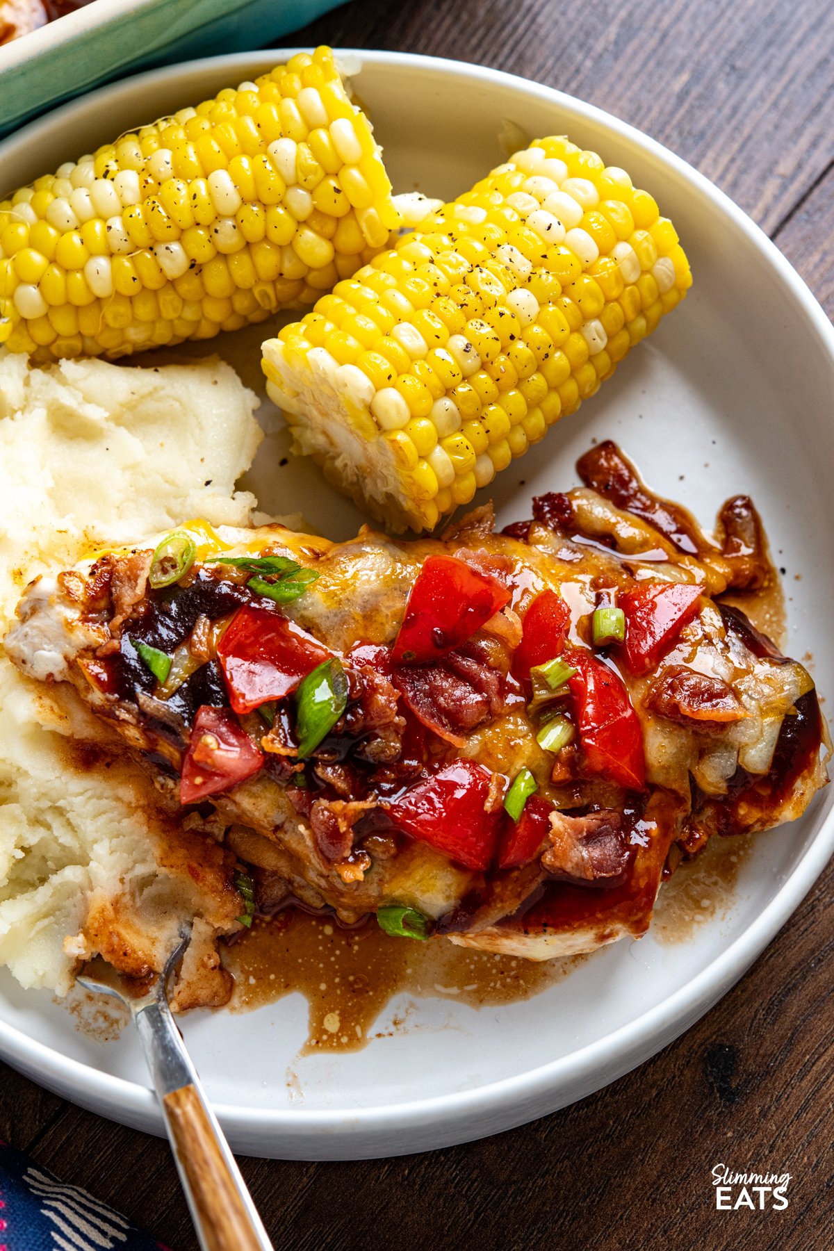 This easy Monterey Chicken is loaded with flavour, featuring tender chicken breasts topped with smoky BBQ sauce, melted cheese, fresh tomatoes and crispy bacon for a fuss-free dinner the whole family will love. close up of white plate of Monterey Chicken served with mashed potatoes and corn on the cob.