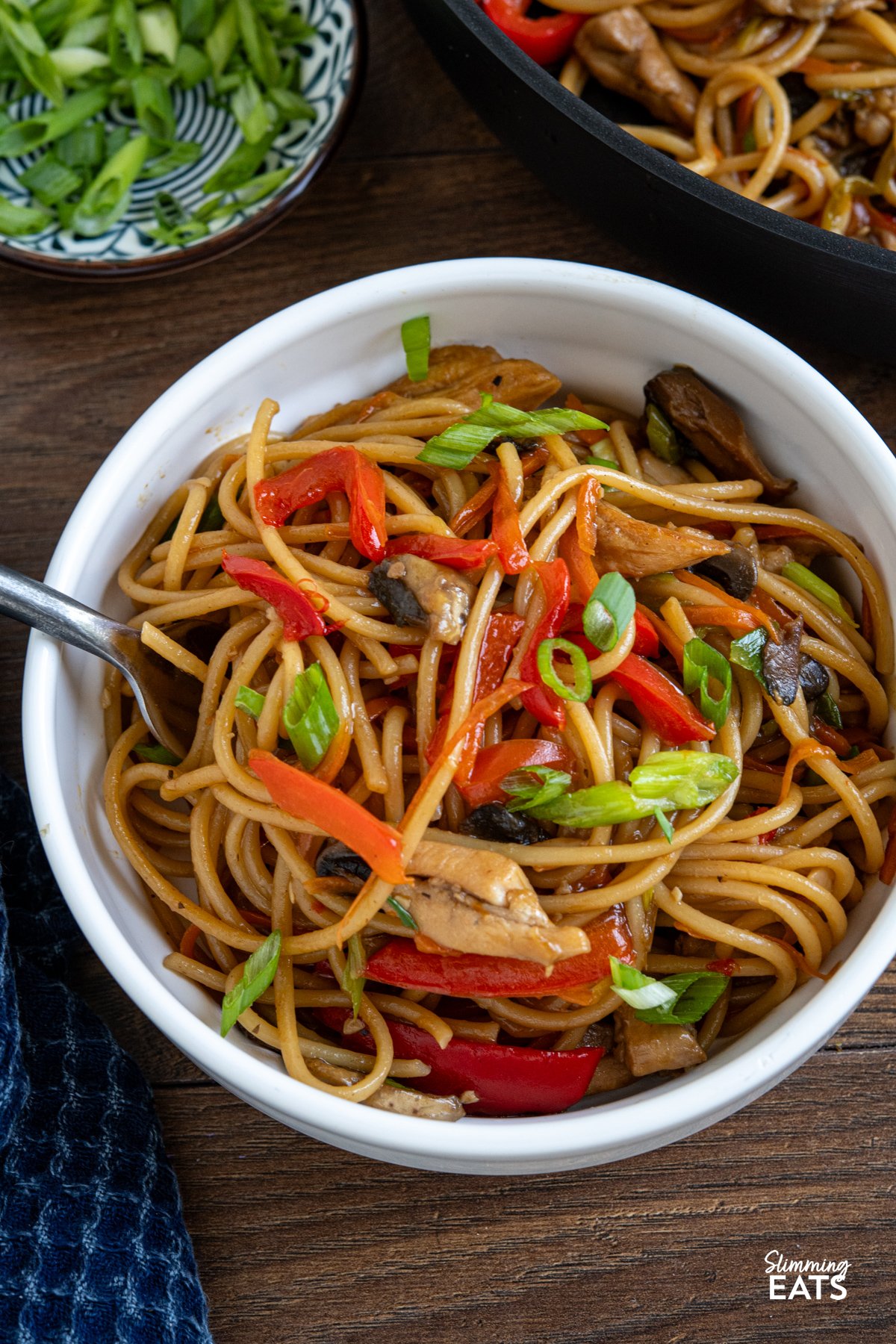 Say hello to your new weeknight favourite: One Pot Asian Garlic Chicken Noodles. This is everything you want from a quick noodle dish: tender chunks of chicken, loads of garlicky flavour, and a glossy, savoury sauce that clings to every strand of spaghetti. Yes, spaghetti! It might not be traditional, but it works perfectly in this one pot recipe, soaking up all those bold, punchy flavours without the fuss of boiling separately. Minimal effort, maximum flavour, and hardly any washing up. close up of serving of asian garlic chicken noodles made with spaghetti in a small white bowl next to pan of noodles.