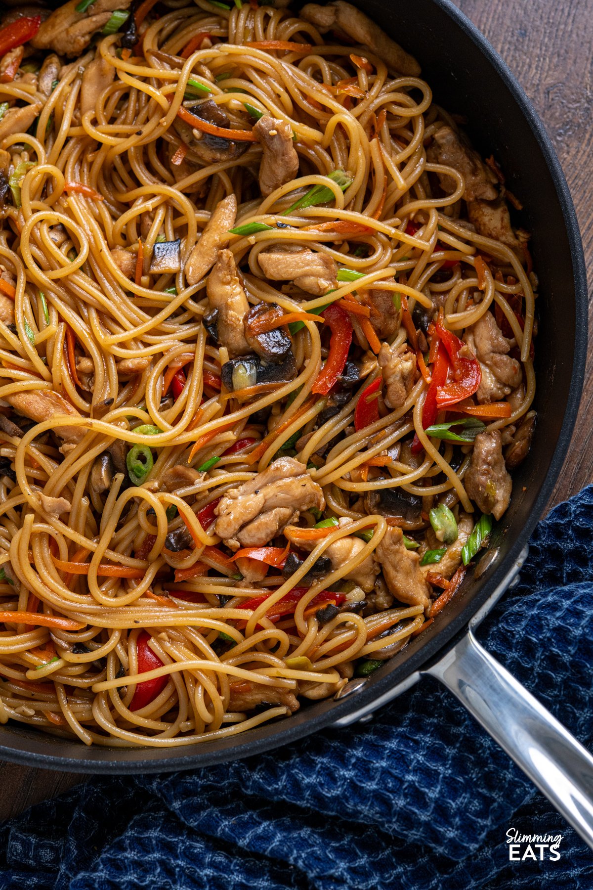 Say hello to your new weeknight favourite: One Pot Asian Garlic Chicken Noodles. This is everything you want from a quick noodle dish: tender chunks of chicken, loads of garlicky flavour, and a glossy, savoury sauce that clings to every strand of spaghetti. Yes, spaghetti! It might not be traditional, but it works perfectly in this one pot recipe, soaking up all those bold, punchy flavours without the fuss of boiling separately. Minimal effort, maximum flavour, and hardly any washing up. close up of one pot garlic chicken noodles made with spaghetti in a all-clad non stick pan next to a blue kitchen towel.