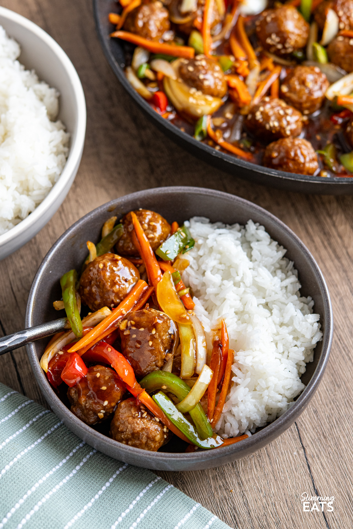 This Easy Asian Meatball Stir Fry is a quick and flavourful weeknight dinner that brings tender meatballs and vibrant vegetables together in a glossy savoury sweet sauce. Easy Asian Meatball Stir Fry in grey bowl with rice, frying pan of meatballs and bowl of rice of visible in background with side of rice