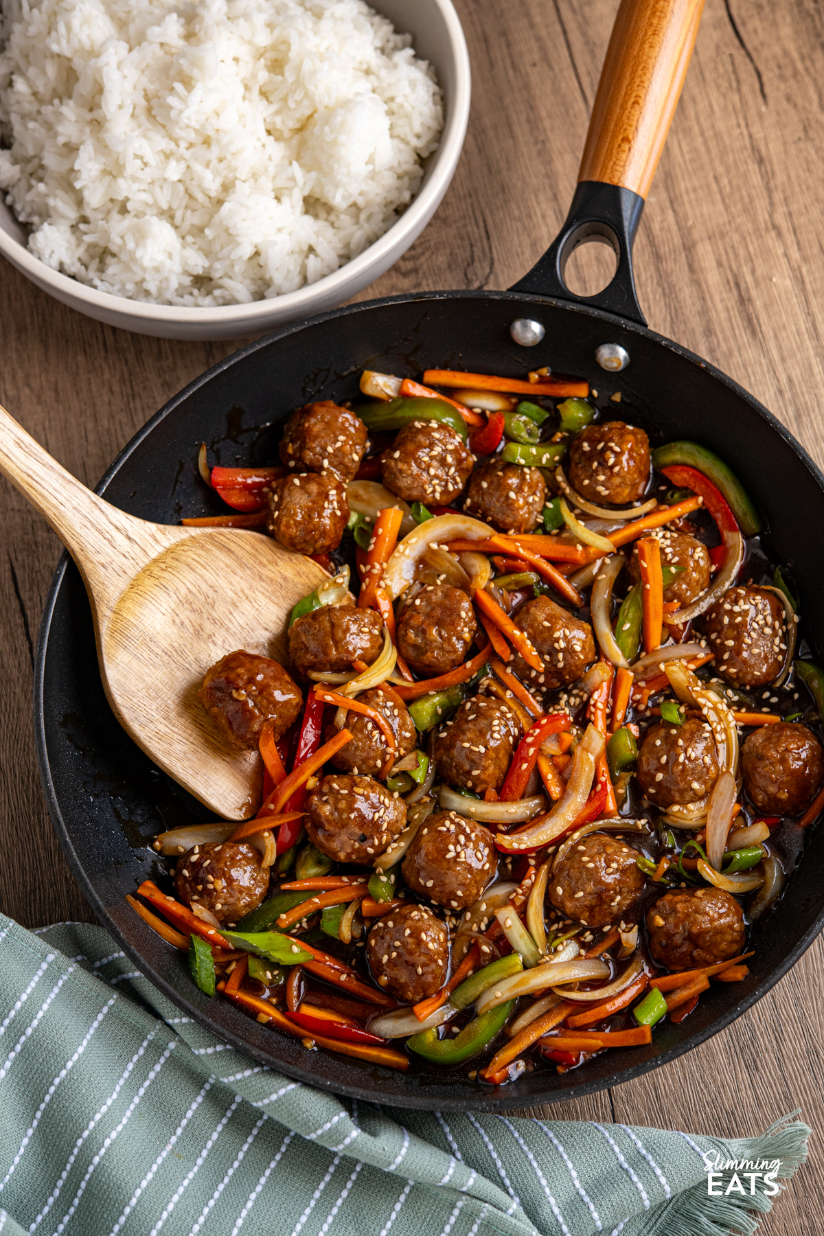 This Easy Asian Meatball Stir Fry is a quick and flavourful weeknight dinner that brings tender meatballs and vibrant vegetables together in a glossy savoury sweet sauce. Easy Asian Meatball Stir Fry in a black non stick frying pan with wooden spoon, rice visible in bowl
