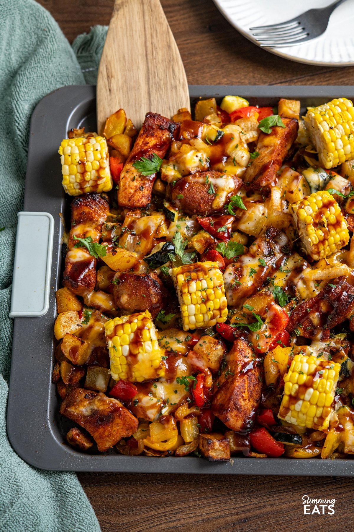 This Sheet Pan BBQ Boneless Pork with Potatoes and Vegetables is an easy, flavour packed dinner. Thick cut pork loin chops roast with yellow potatoes, corn, bell pepper, and zucchini, then get finished with a drizzle of BBQ sauce and a sprinkle of cheese for a smoky, savoury traybake you can bring straight to the table. Cheesy BBQ Boneless Pork Chops and Veggies on a sheet pan with wooden spatula, plate with fork in background.