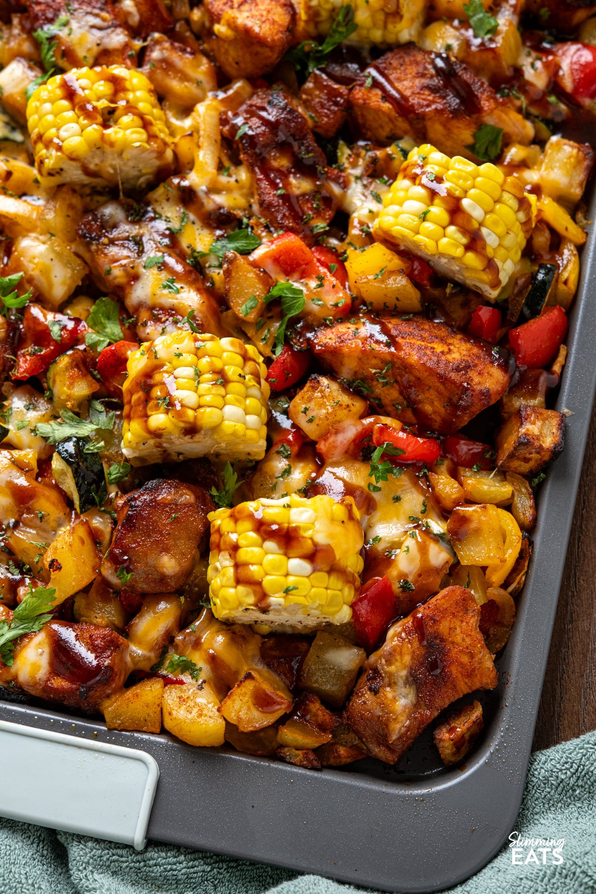 This Sheet Pan BBQ Boneless Pork with Potatoes and Vegetables is an easy, flavour packed dinner. Thick cut pork loin chops roast with yellow potatoes, corn, bell pepper, and zucchini, then get finished with a drizzle of BBQ sauce and a sprinkle of cheese for a smoky, savoury traybake you can bring straight to the table. close up of Cheesy BBQ Boneless Pork Chops and Veggies on a sheet pan tray