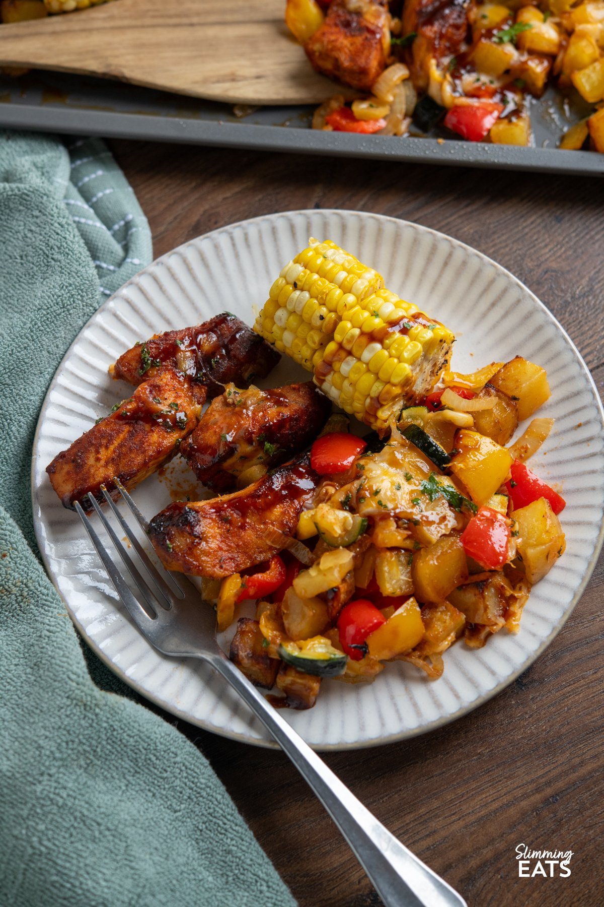 This Sheet Pan BBQ Boneless Pork with Potatoes and Vegetables is an easy, flavour packed dinner. Thick cut pork loin chops roast with yellow potatoes, corn, bell pepper, and zucchini, then get finished with a drizzle of BBQ sauce and a sprinkle of cheese for a smoky, savoury traybake you can bring straight to the table. close up of Cheesy BBQ Boneless Pork Chops and Veggies on a white plate with fork, sheet pan visible in background and green gray napkin to the left