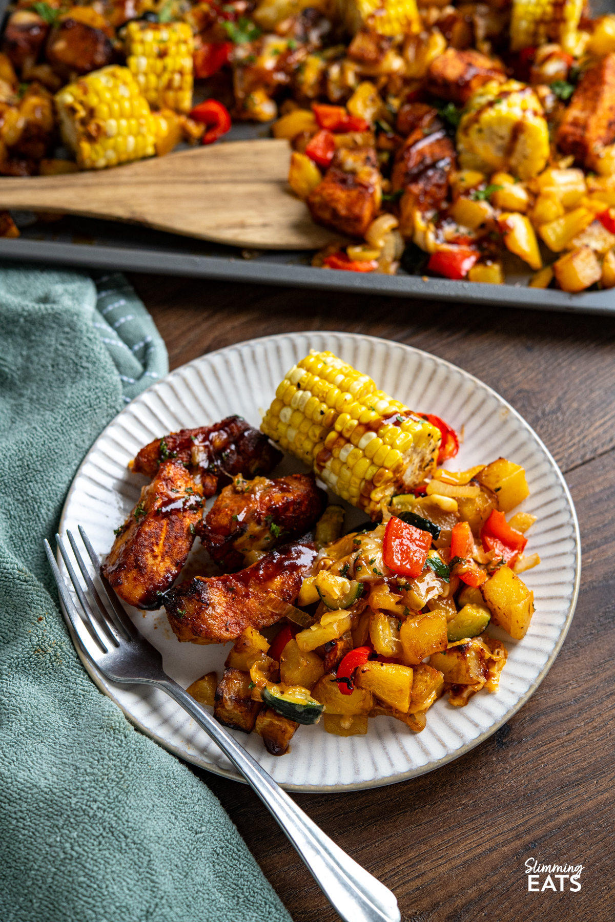 This Sheet Pan BBQ Boneless Pork with Potatoes and Vegetables is an easy, flavour packed dinner. Thick cut pork loin chops roast with yellow potatoes, corn, bell pepper, and zucchini, then get finished with a drizzle of BBQ sauce and a sprinkle of cheese for a smoky, savoury traybake you can bring straight to the table. Cheesy BBQ Boneless Pork Chops and Veggies on a white plate with fork, sheet pan visible in background and green gray napkin to the left