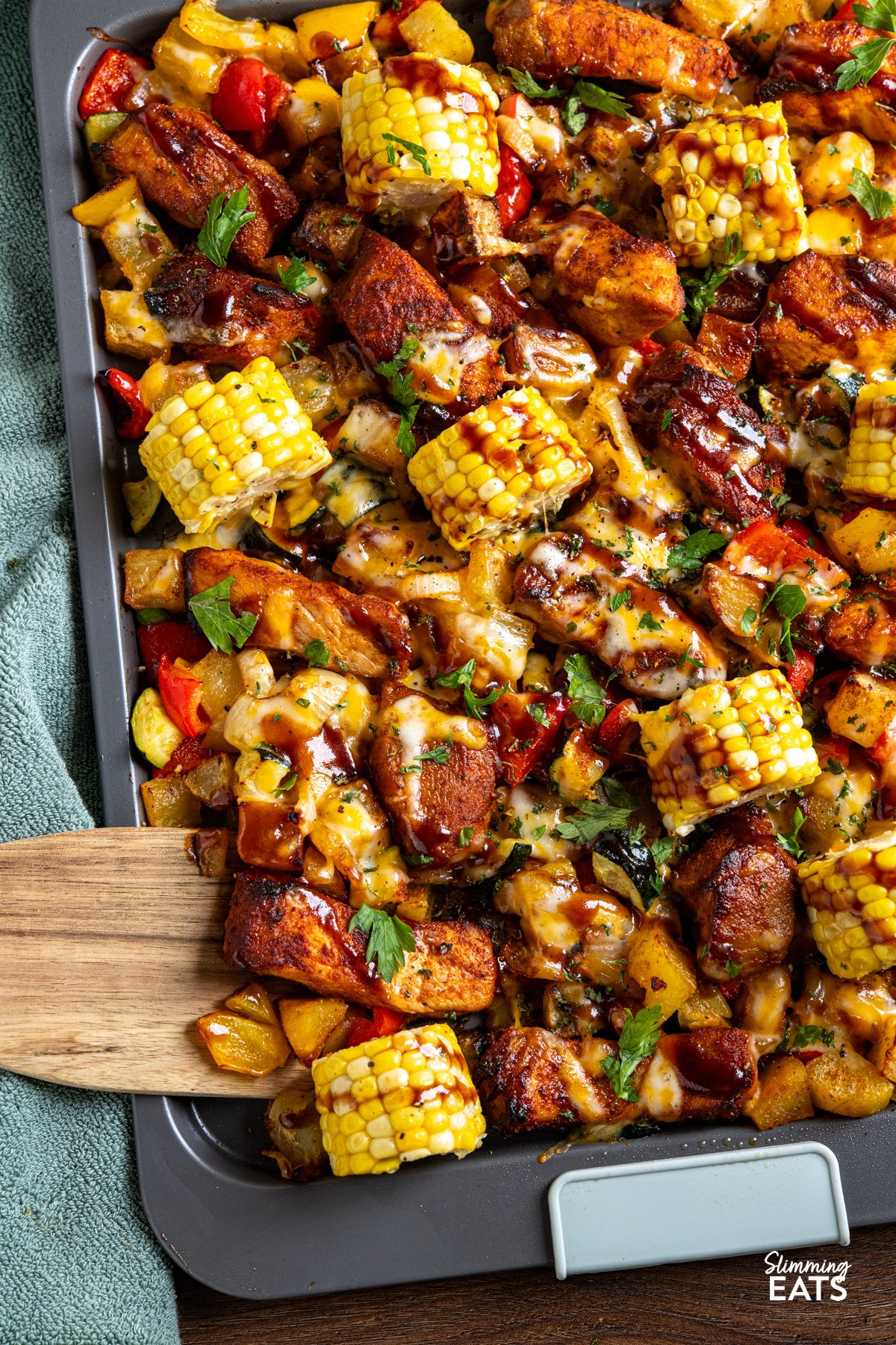 This Sheet Pan BBQ Boneless Pork with Potatoes and Vegetables is an easy, flavour packed dinner. Thick cut pork loin chops roast with yellow potatoes, corn, bell pepper, and zucchini, then get finished with a drizzle of BBQ sauce and a sprinkle of cheese for a smoky, savoury traybake you can bring straight to the table. close up of Cheesy BBQ Boneless Pork Chops and Veggies on a sheet pan with wooden spatula