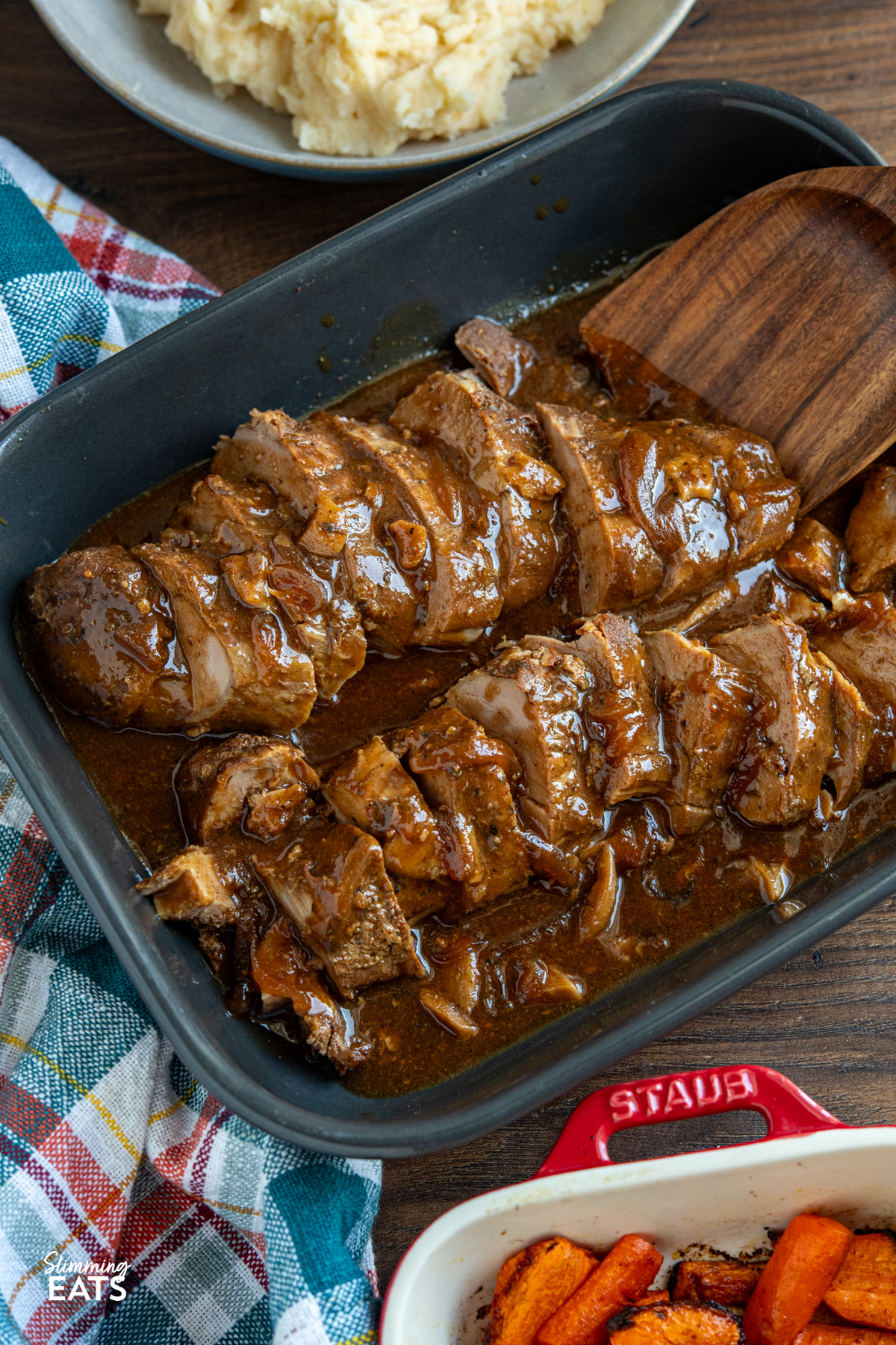 This Slow Cooker Honey Soy Balsamic Pork Tenderloin is an easy set-and-forget dinner. Simply whisk together balsamic vinegar, soy sauce, honey and garlic with a touch of seasoning, pour it over the pork and let the slow cooker work its magic. In just a few hours, you will have tender pork coated in a sticky, sweet and savoury sauce. slow cooker honey soy balsamic pork tenderloin sliced in a grey dish with the sauce, wooden spatula is in the dish ready to serve up some slices, sides of mashed potato and roasted carrots visible.