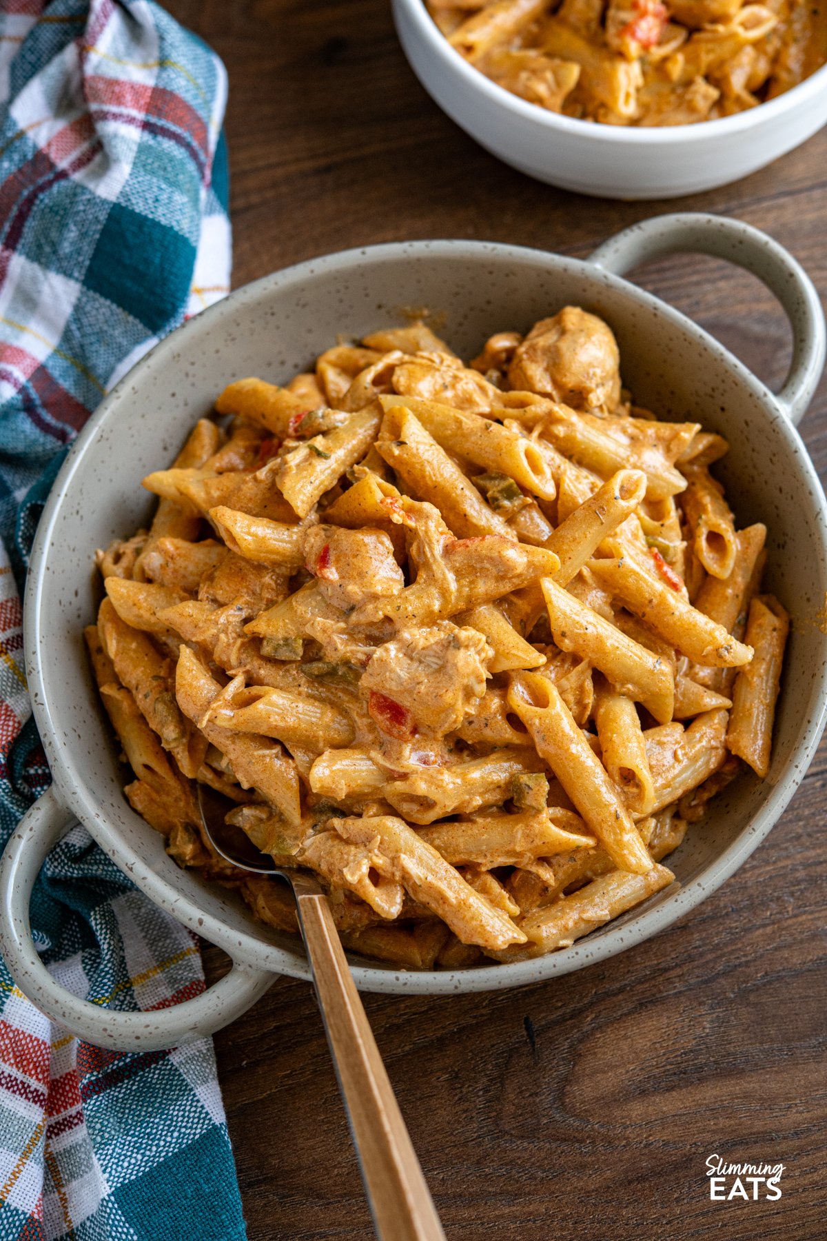 Slow Cooker Creamy Cajun Chicken Pasta is an easy, all-in-one meal with tender chicken, a light creamy sauce, and pasta cooked right in the slow cooker. Slow Cooker Creamy Cajun Chicken Pasta served in a double handled grey brown ceramic dish, with a bowl of pasta in the background. A blue, red, white, and yellow chequered kitchen towel is placed to the left.