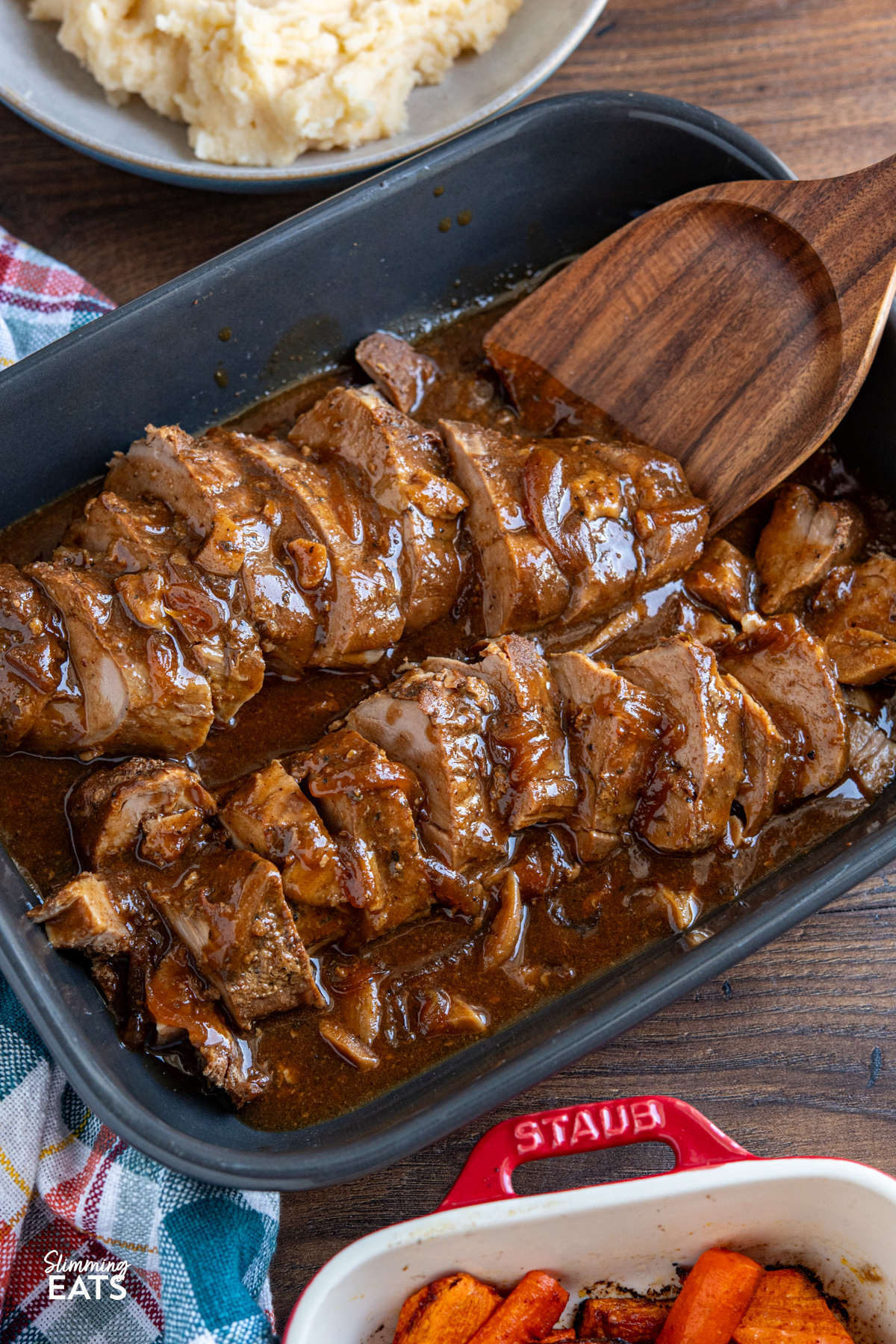 slow cooker honey soy balsamic pork tenderloin sliced in a grey dish with the sauce, wooden spatula is in the dish ready to serve up some slices, sides of mashed potato and roasted carrots visible. 