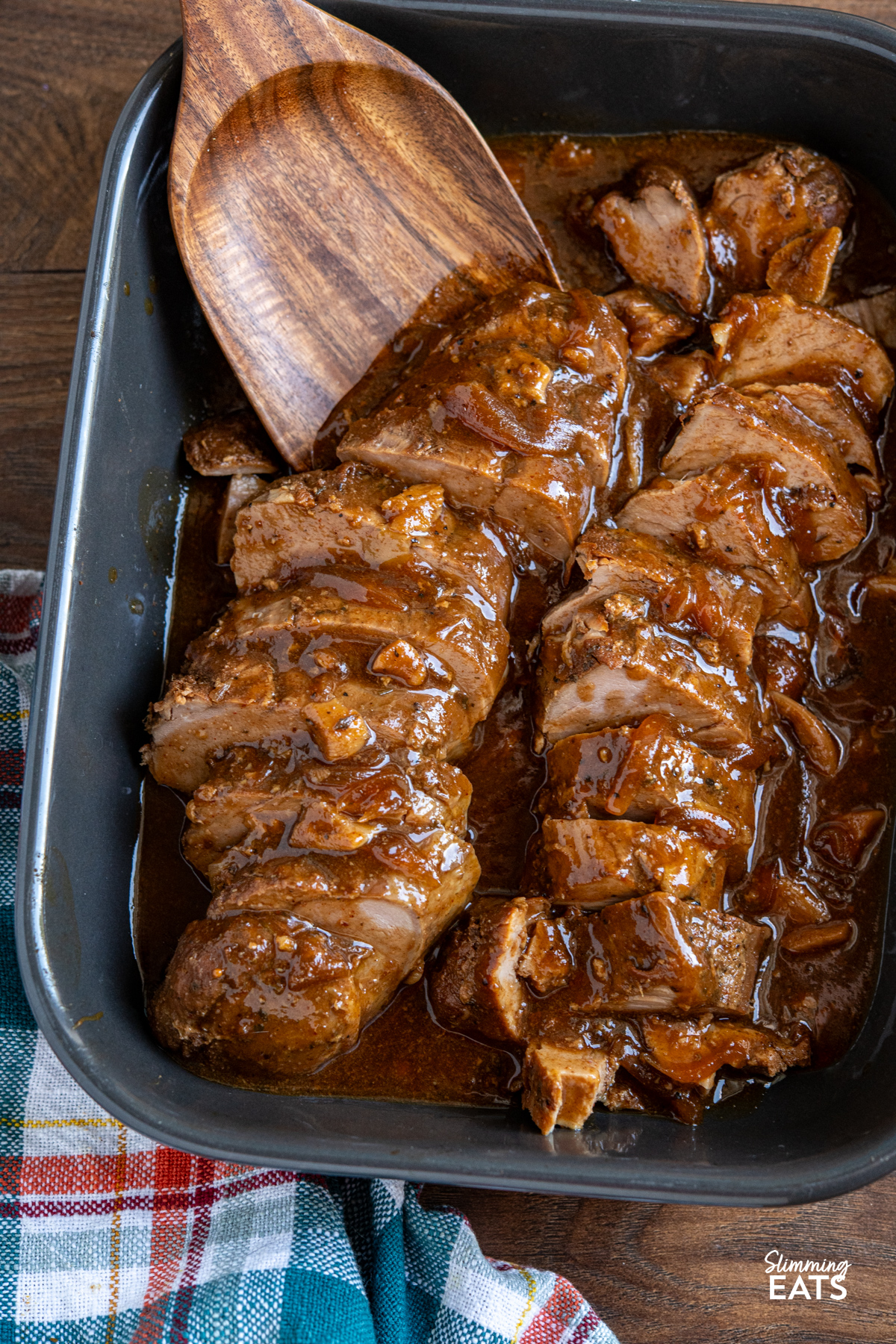 slow cooker honey soy balsamic pork tenderloin sliced in a grey dish with the sauce, wooden spatula is in the dish ready to serve up some slices