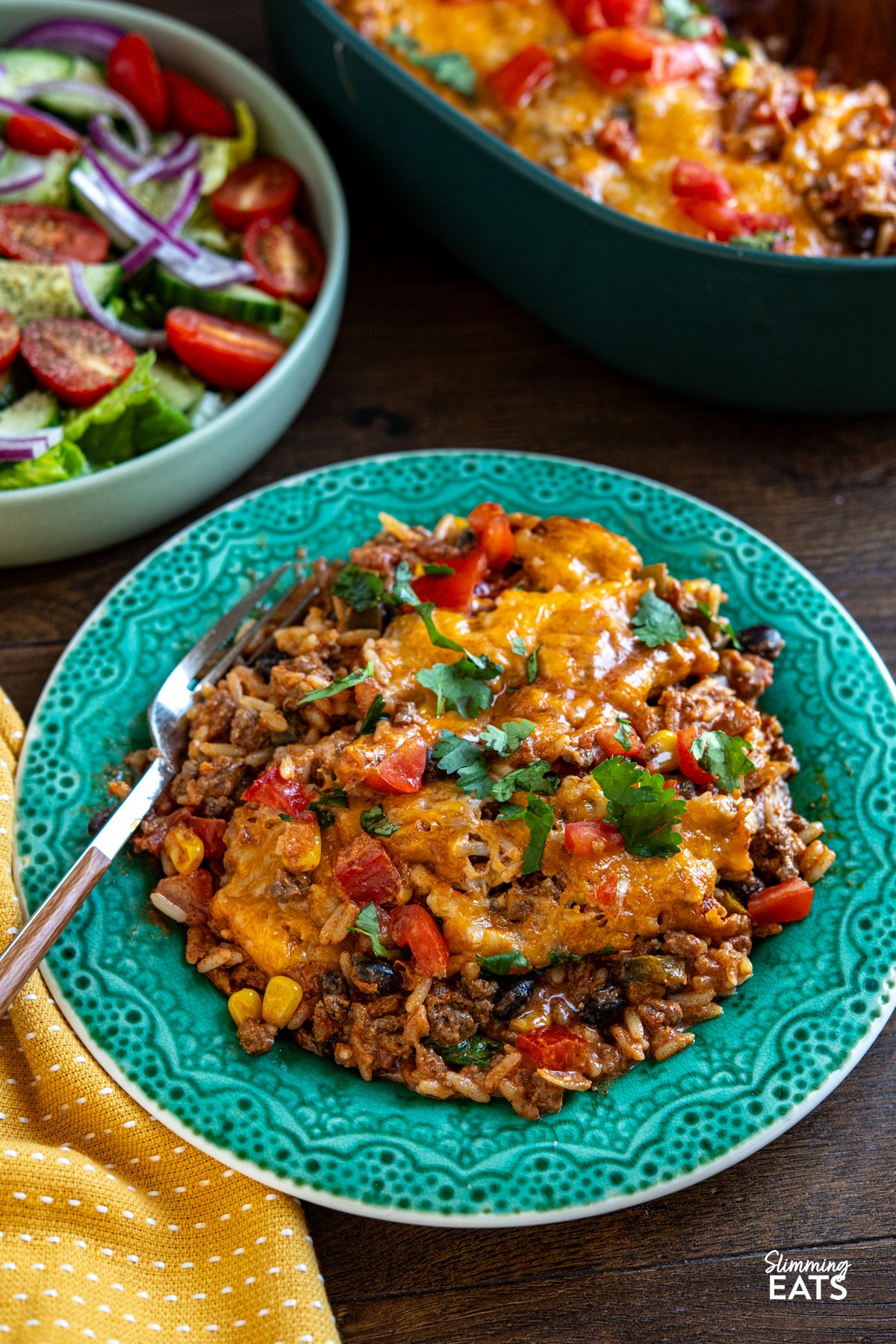 Enjoy all the flavours of a burrito in this easy Cheesy Beef Burrito Rice Bake packed with seasoned ground beef, fluffy rice, sweetcorn, black beans, and colourful peppers, every bite is full of Tex-Mex flair. serving of Cheesy Beef Burrito Rice Bake on a dark turquoise patterned plate with bowl of salad visible