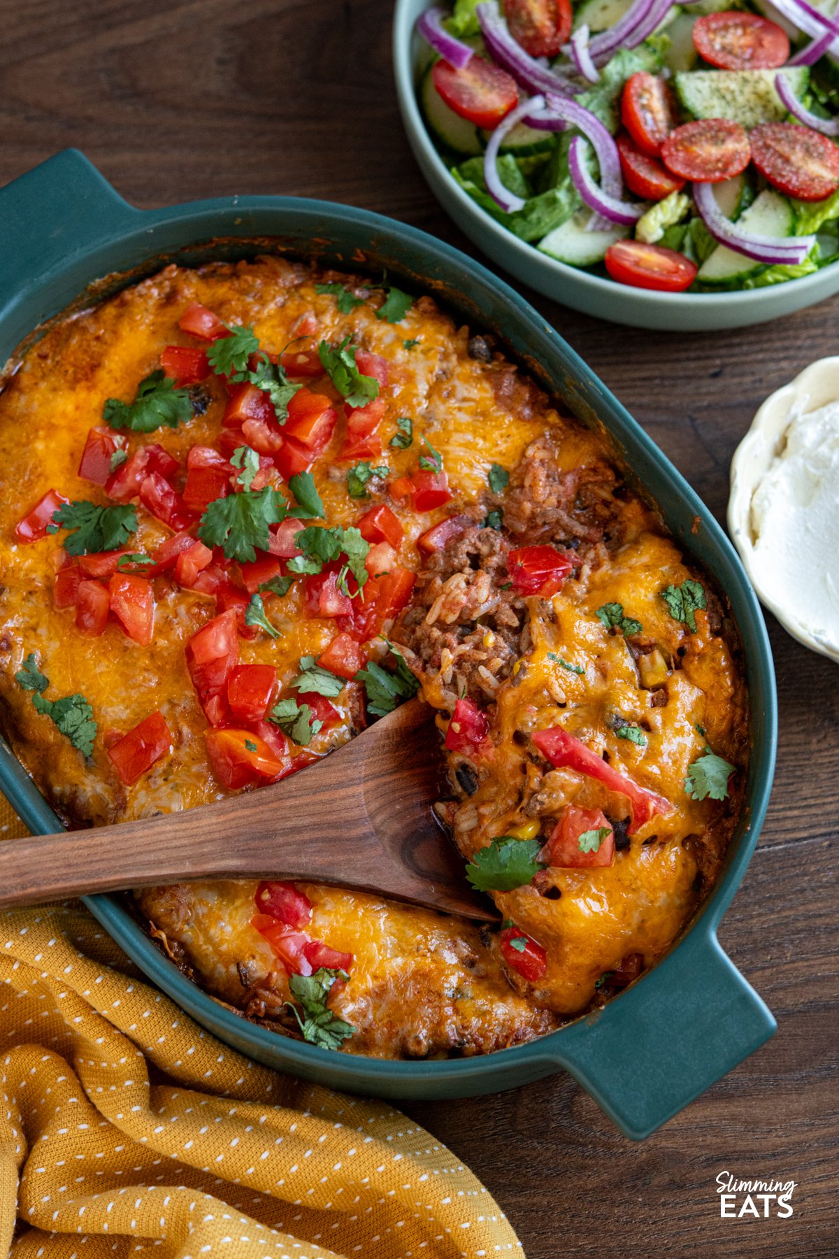 Enjoy all the flavours of a burrito in this easy Cheesy Beef Burrito Rice Bake packed with seasoned ground beef, fluffy rice, sweetcorn, black beans, and colourful peppers, every bite is full of Tex-Mex flair. Cheesy Beef Burrito Rice Bake in dark green oven dish with bowl of salad and soured cream to the right.