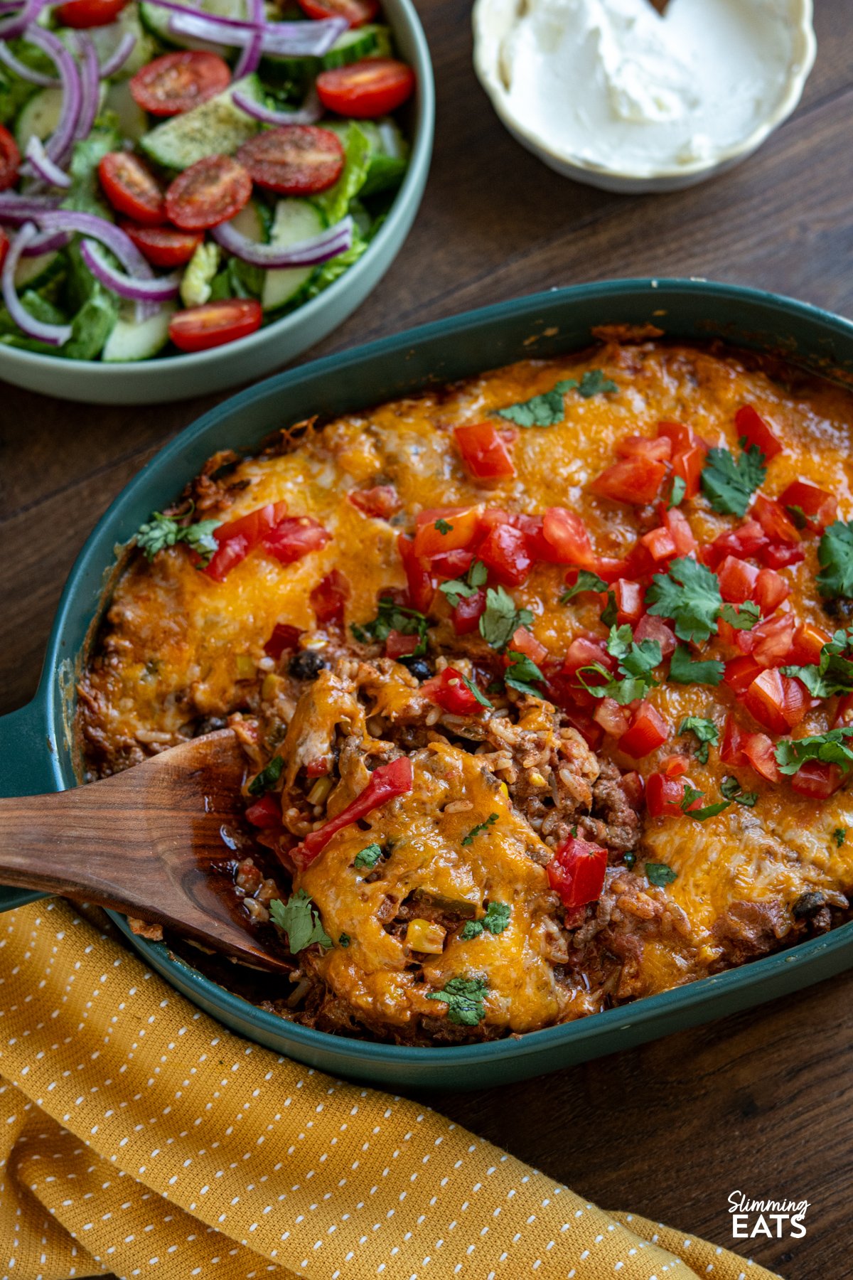 Enjoy all the flavours of a burrito in this easy Cheesy Beef Burrito Rice Bake packed with seasoned ground beef, fluffy rice, sweetcorn, black beans, and colourful peppers, every bite is full of Tex-Mex flair. Cheesy Beef Burrito Rice Bake in dark green oven dish with bowl of salad and soured cream above