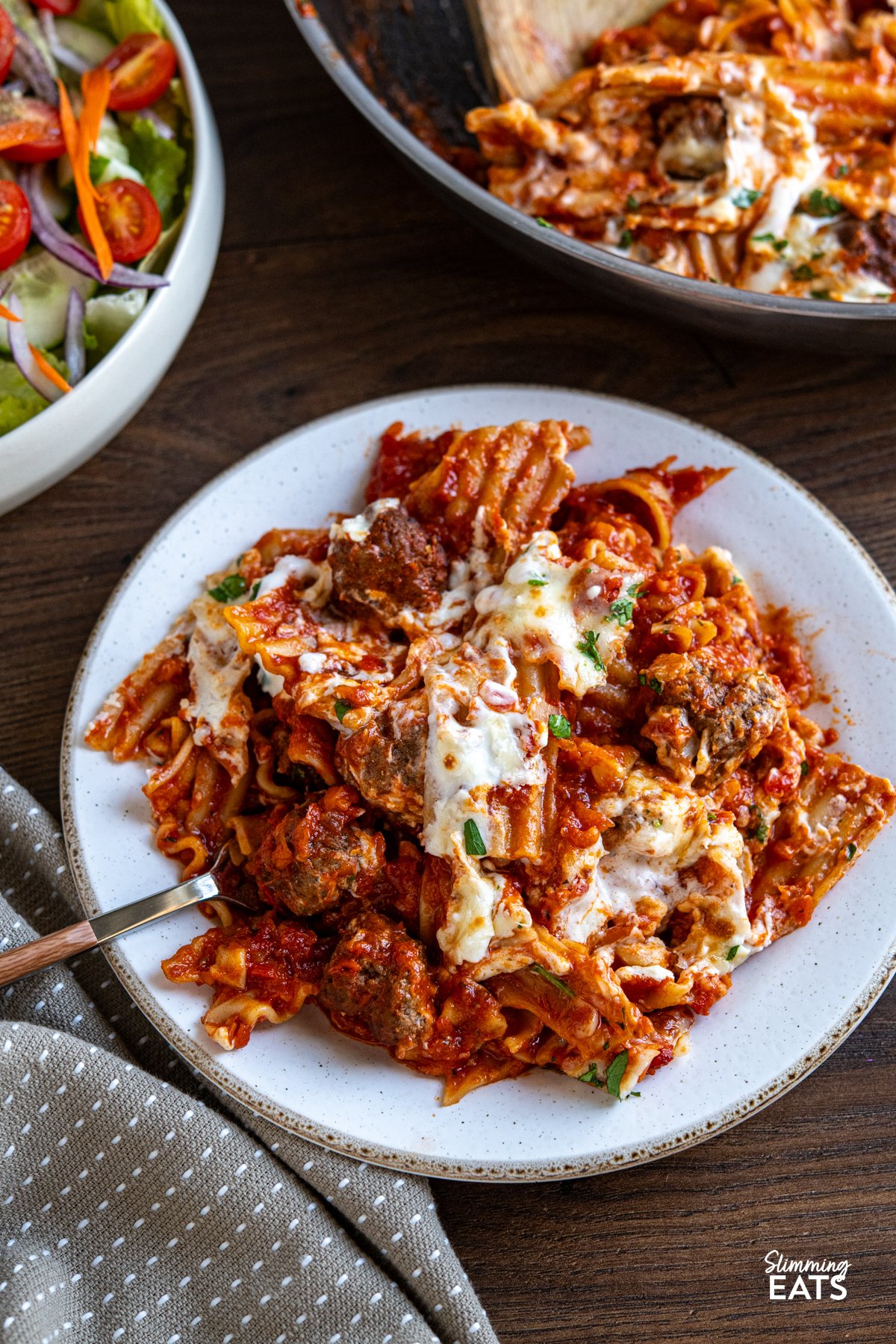 This One Pan Lasagna Meatball Skillet combines tender homemade meatballs, broken lasagne sheets, and a rich tomato sauce with a lighter creamy béchamel for a comforting, fuss free family friendly dinner. serving of lasagna Meatballs on white plate with a beige rim, fork placed into the pasta.