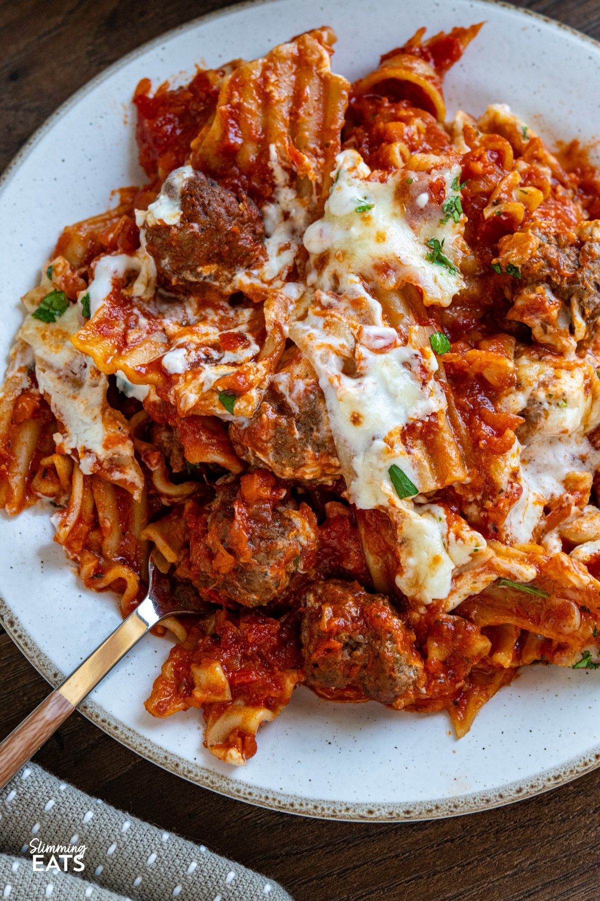 This One Pan Lasagna Meatball Skillet combines tender homemade meatballs, broken lasagne sheets, and a rich tomato sauce with a lighter creamy béchamel for a comforting, fuss free family friendly dinner. close up of serving of lasagna Meatballs on white plate with a beige rim, fork placed into the pasta.