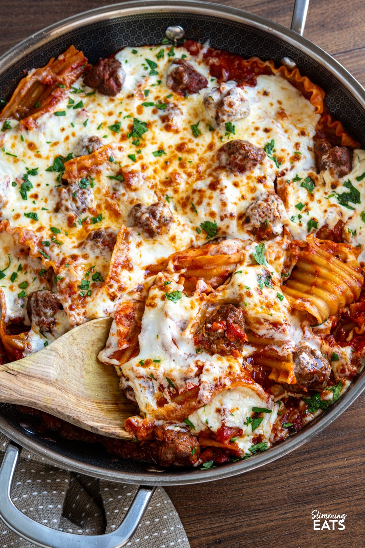 This One Pan Lasagna Meatball Skillet combines tender homemade meatballs, broken lasagne sheets, and a rich tomato sauce with a lighter creamy béchamel for a comforting, fuss free family friendly dinner. close up of spatula in One Pan Lasagna Meatballs in double handled non stick skillet