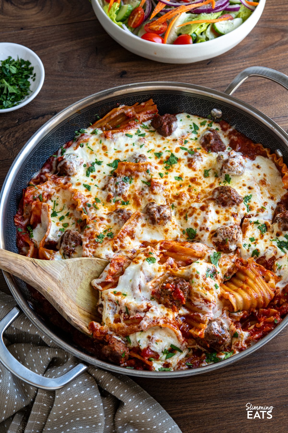 This One Pan Lasagna Meatball Skillet combines tender homemade meatballs, broken lasagne sheets, and a rich tomato sauce with a lighter creamy béchamel for a comforting, fuss free family friendly dinner. One Pan Lasagna Meatballs in double handled non stick skillet with bowl of mixed salad and small bowl of parsley nearby