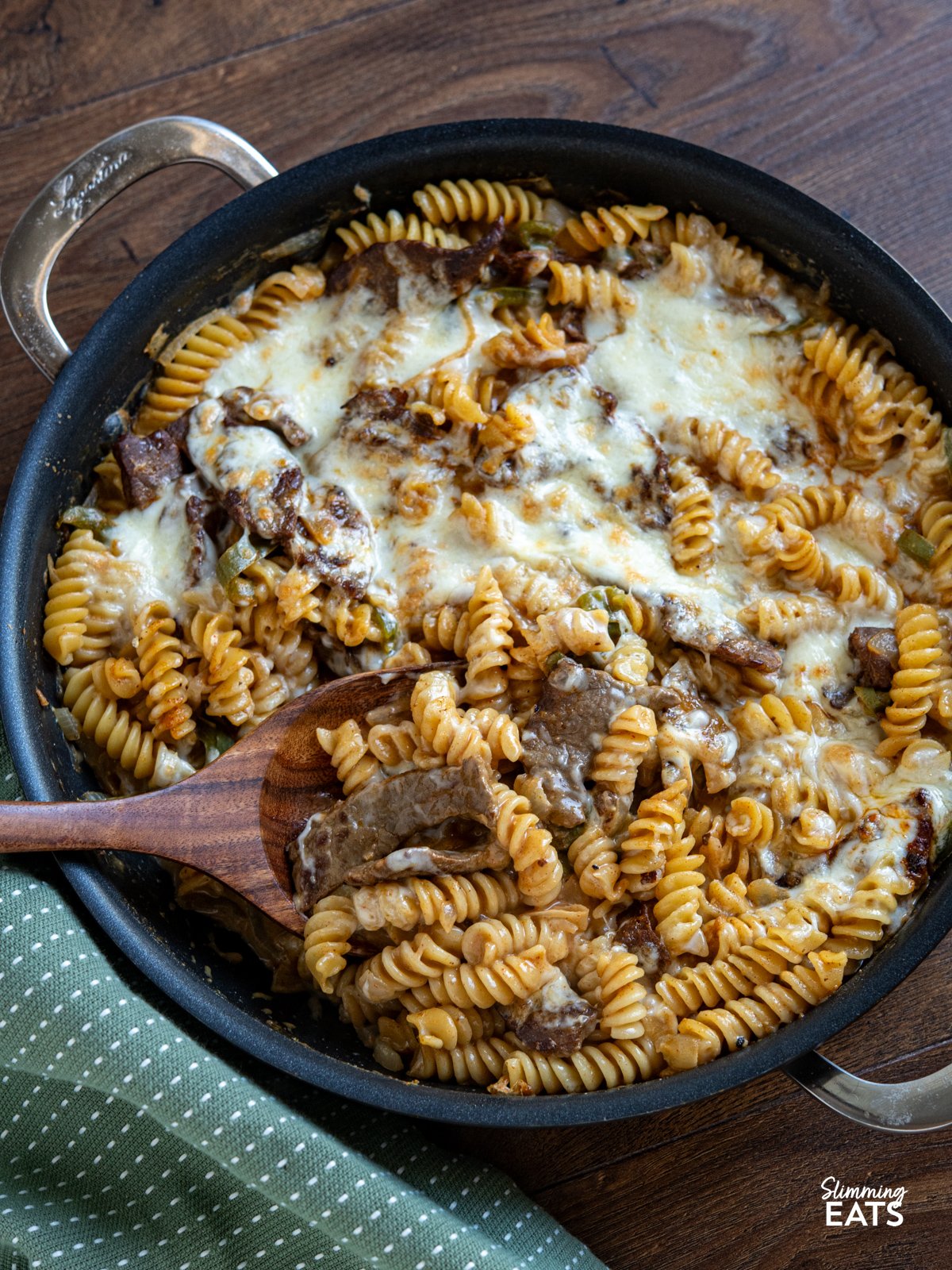 This One-Pot Philly Cheesesteak Pasta is a creamy, cheesy twist on the classic sandwich, with tender beef, peppers, and onions all cooked together for an easy, comforting meal. One Pot Philly Cheesesteak Pasta in double handled non stick pan with wooden spatula