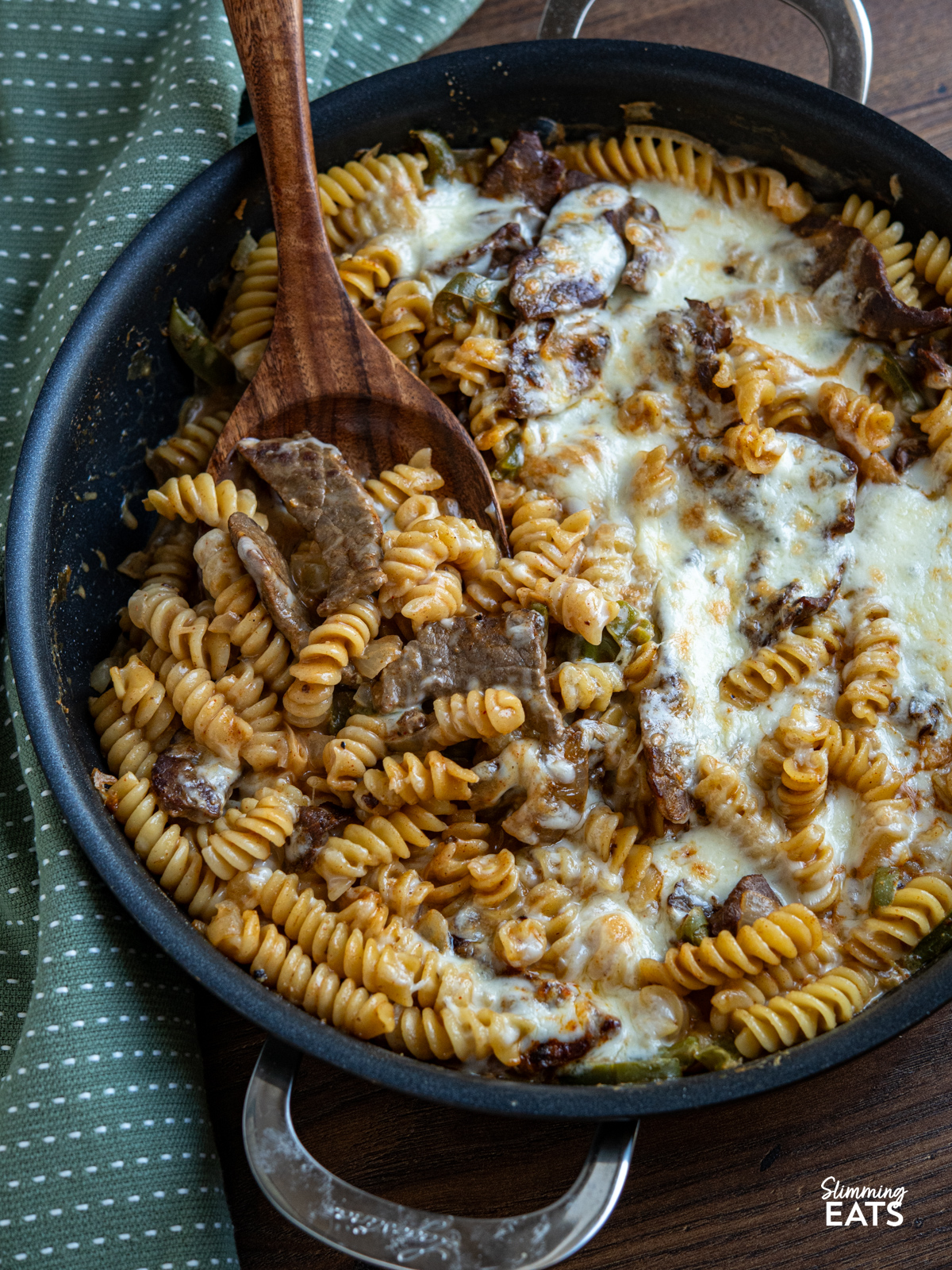 This One Pot Philly Cheesesteak Pasta is a creamy, cheesy twist on the classic sandwich, with tender beef, peppers, and onions all cooked together for an easy, comforting meal. close up of One Pot Philly Cheesesteak Pasta in a double handled non stick pan with wooden spatula