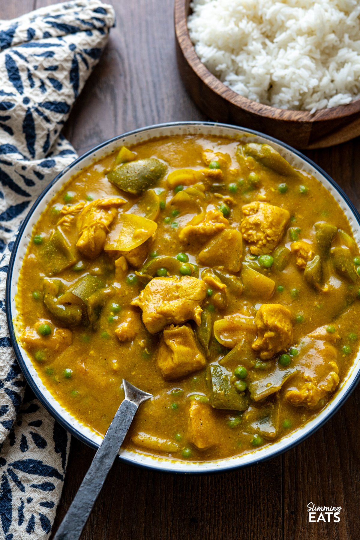 Slow Cooker Chinese Chicken Curry in a large bowl with navy rim, serving spoon placed in bowl, wooden bowl with rice visible