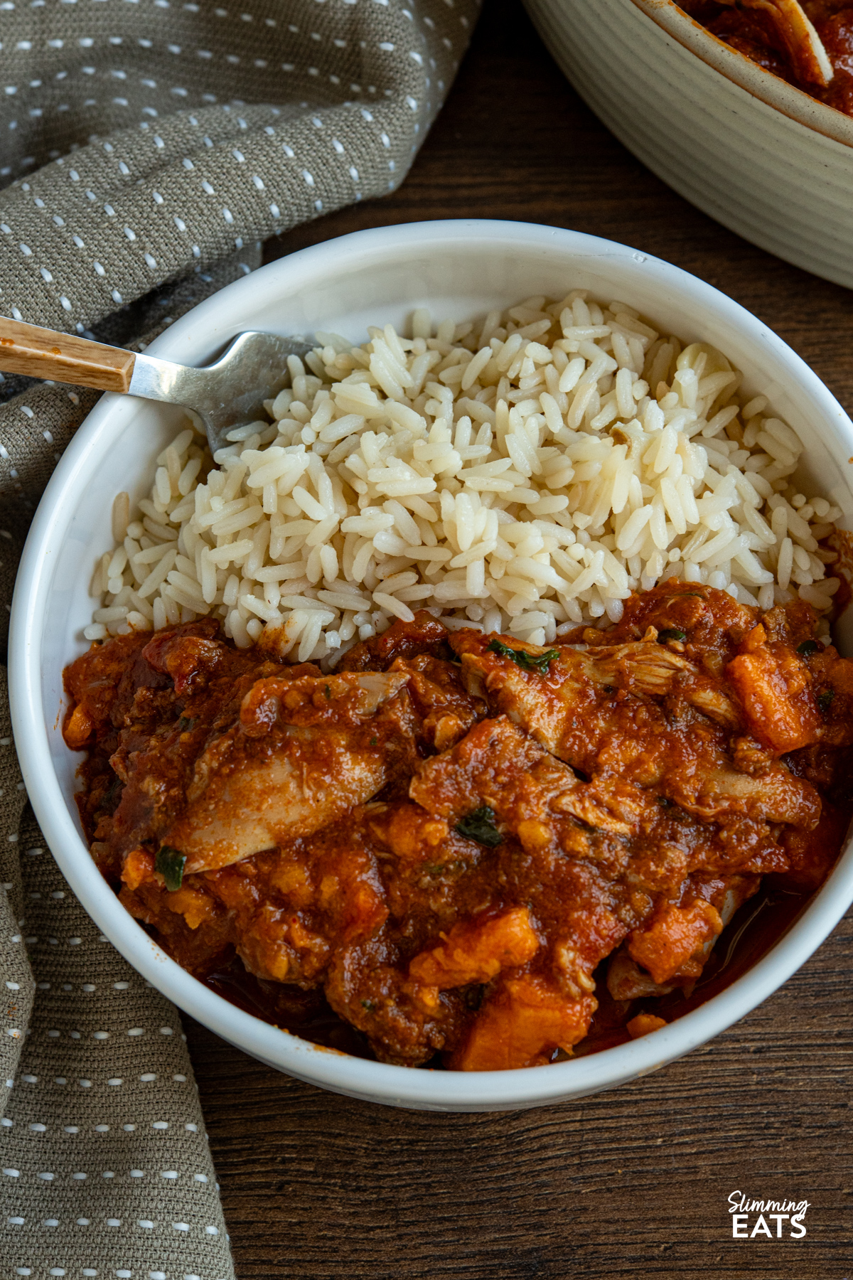 This Slow Cooker Chicken, Chorizo and Sweet Potato Stew is a hearty, flavour-packed meal that brings comfort and richness to any table. close up of Slow Cooker Chicken Chorizo Sweet Potato Stew serving in a white bowl with long grain rice, ceramic double handled dish with the remaining stew visible above.