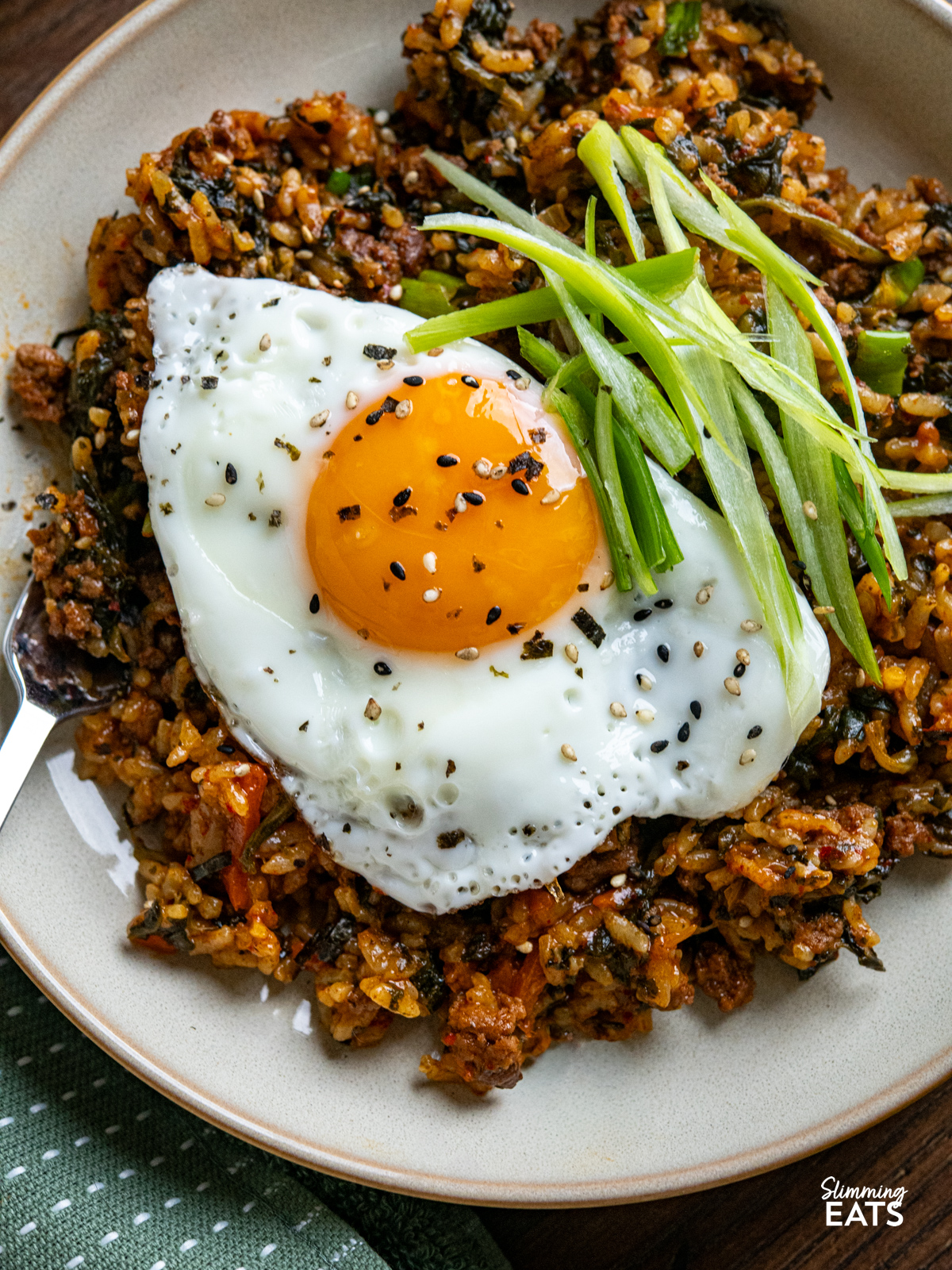 Enjoy the rich, savoury comfort of this Slow Cooker Korean Beef with Spinach and Rice. Ground beef simmers with garlic, ginger, and a sauce where gochujang is the star, alongside spinach and rice for a complete, flavour packed meal. close up of serving of Slow Cooker Korean Beef Spinach Rice in a beige bowl topped with fried egg, sliced green onions, sesame seed nori seasoning
