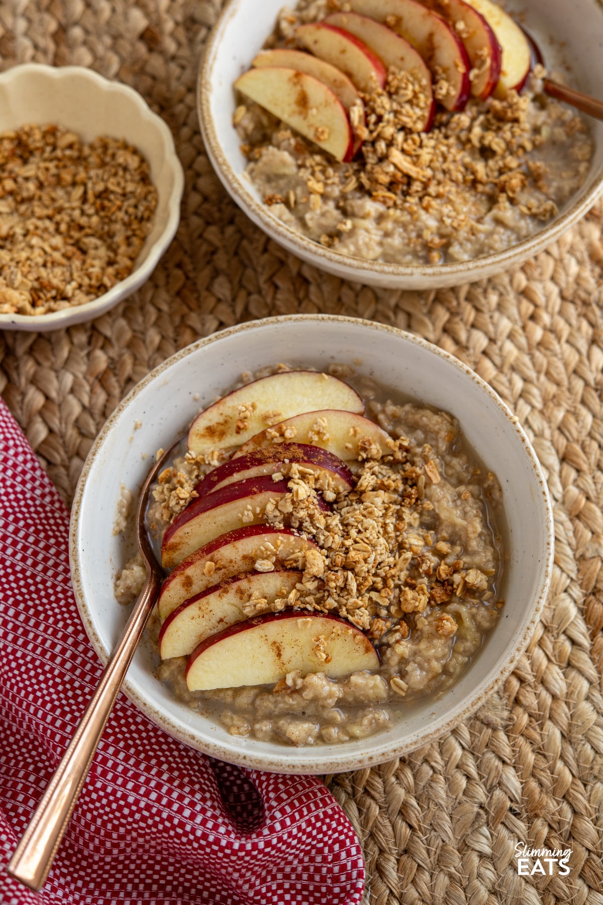 Start your day with this simple and comforting Apple Cinnamon Oatmeal bowl, packed with warming spices, crisp apple and low sugar granola topping. two bowls of apple cinnamon oatmeal with a small bowl of low sugar granola to the side for topping.