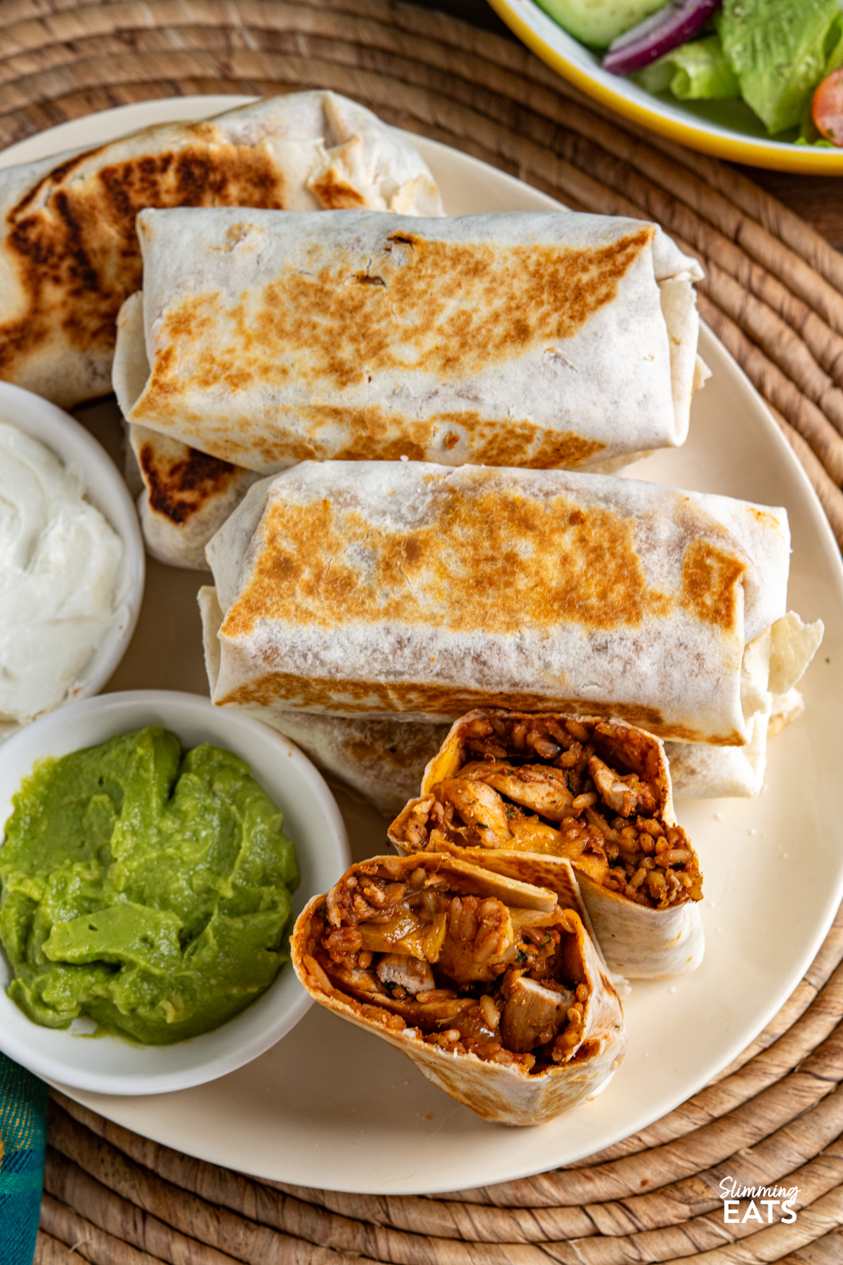 One Pan Chicken Rice Burritos on a oval cream plate with soured cream and guacamole, bowl of salad on side