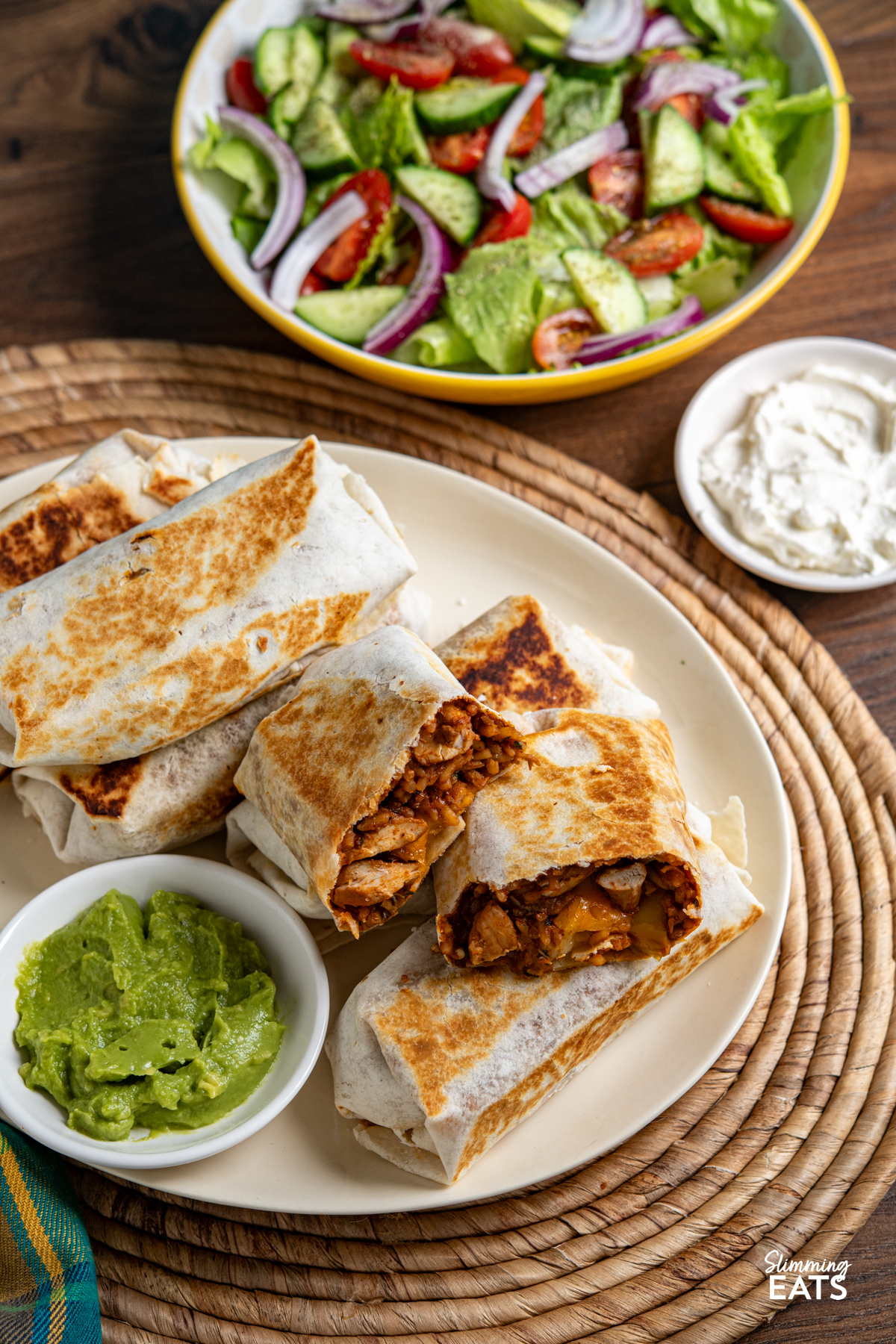 These one pan chicken rice burritos combine tender chicken, peppers, pinto beans and melted cheese for a quick, flavour-packed dinner. One Pan Chicken Rice Burritos on a cream over plate with side of guacamole, bowl of soured cream and salad on the side.