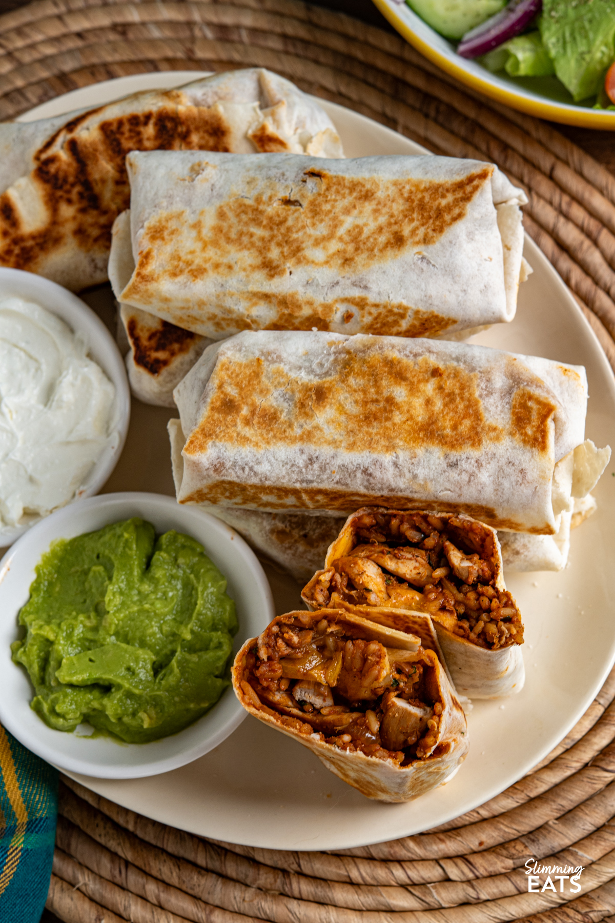These one pan chicken rice burritos combine tender chicken, peppers, pinto beans and melted cheese for a quick, flavour-packed dinner. One Pan Chicken Rice Burritos on a oval cream plate with soured cream and guacamole, bowl of salad on side