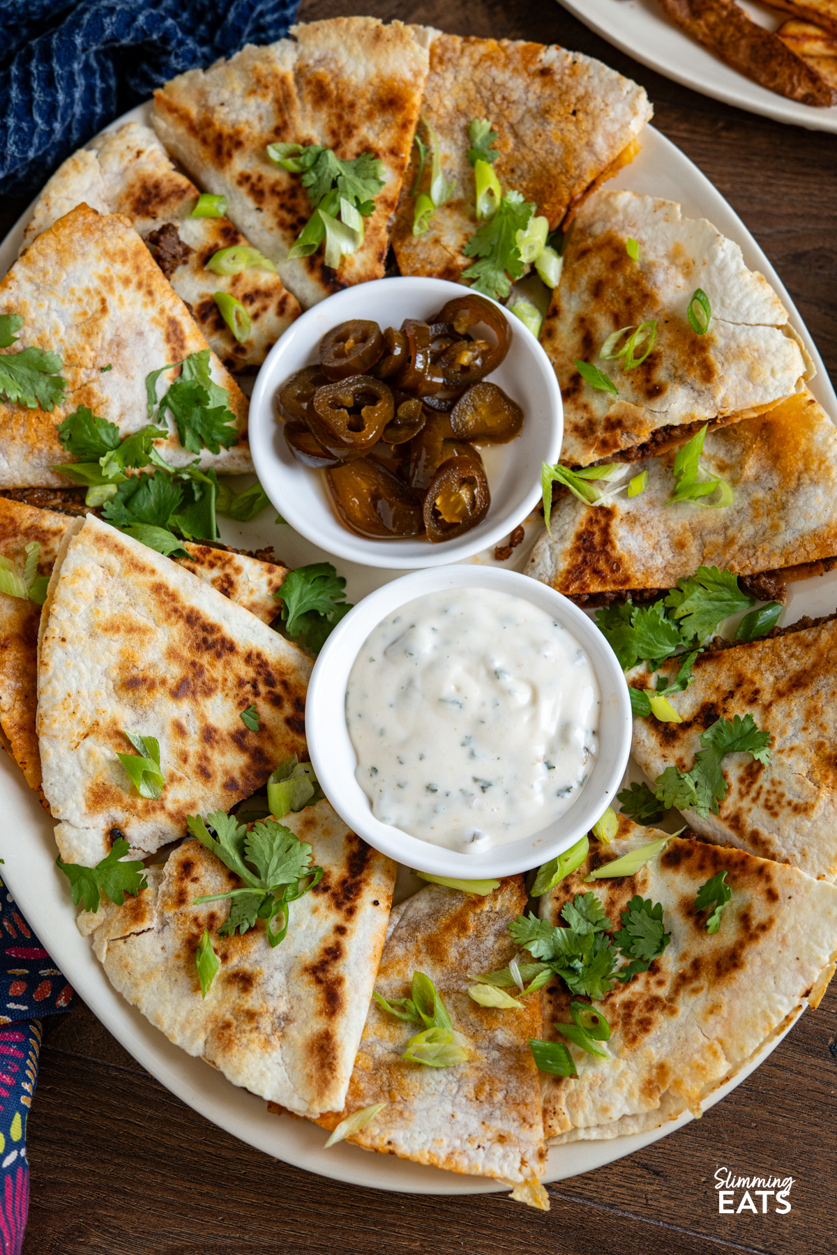 close up of Cheesy BBQ Beef Jalapeno Quesadillas triangles on a oval plate with a small bowl of pickled jalapenos and creamy dip.