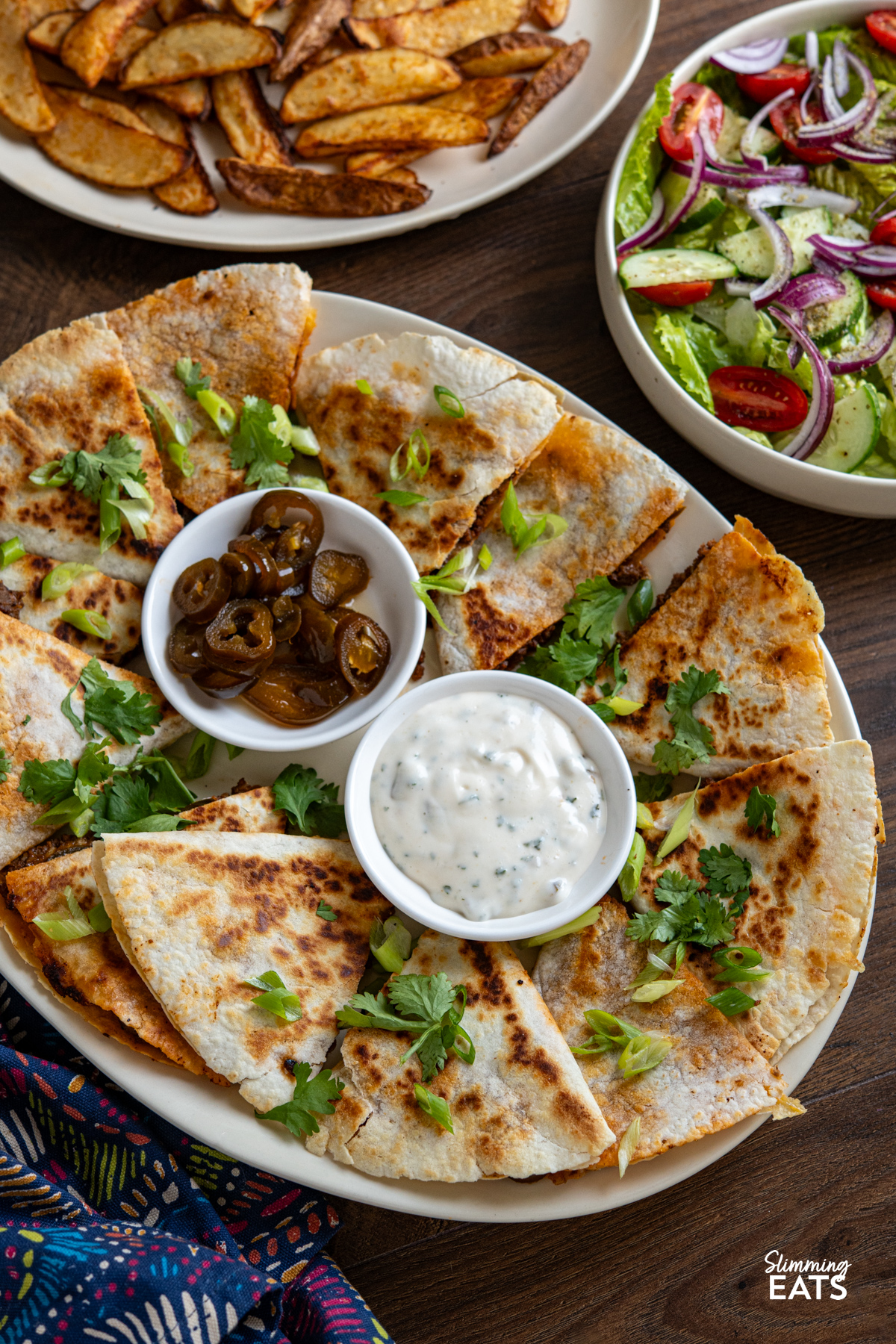 Cheesy BBQ Beef Jalapeno Quesadillas triangles on a oval plate with a small bowl of pickled jalapenos and creamy dip, surrounded by plate or wedges and bowl of mixed salad.