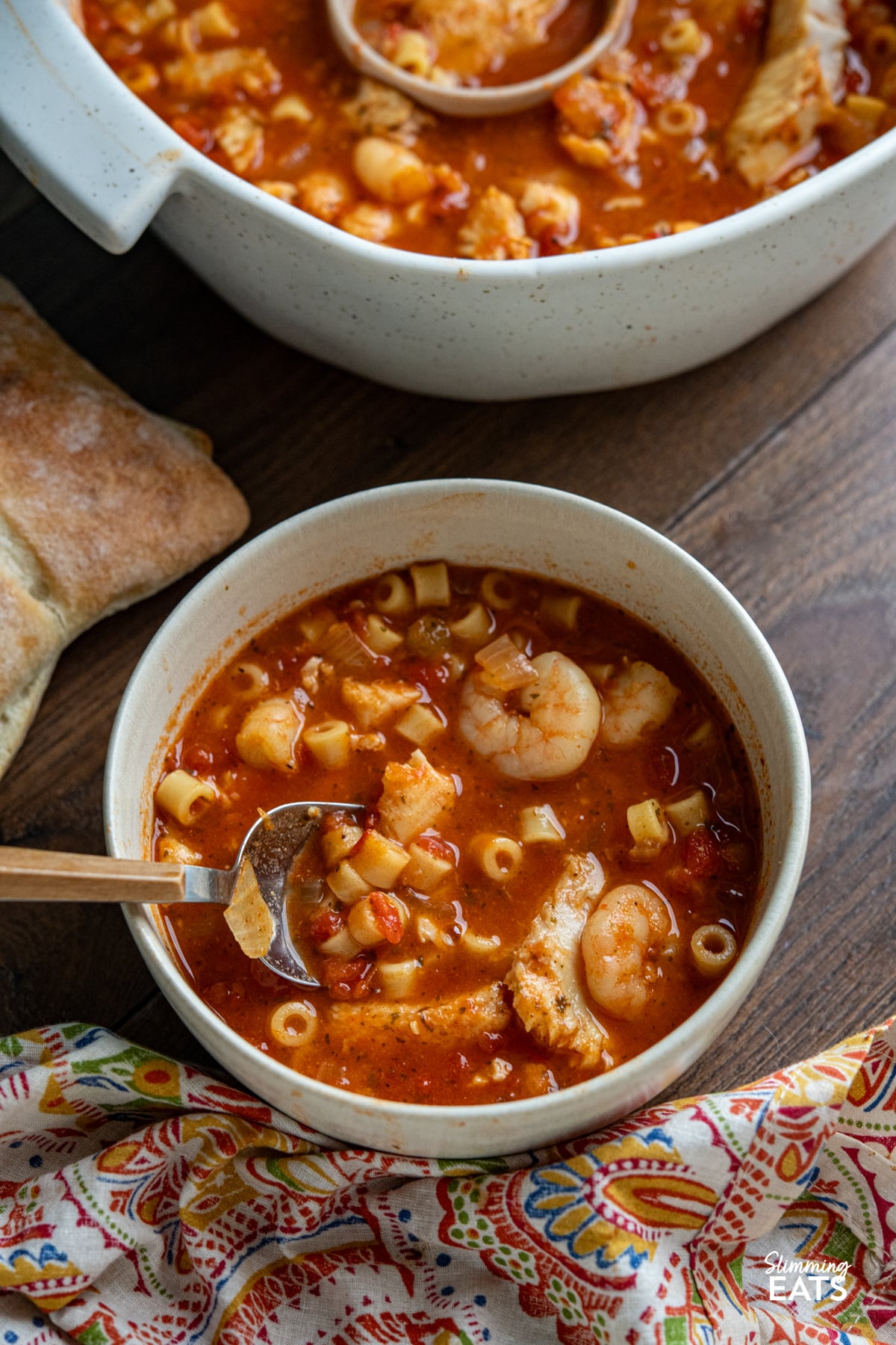 A rich and hearty Mediterranean-inspired seafood pasta soup made with monkfish, shrimp and tender ditalini in a deeply flavourful tomato broth. serving of Fish and Pasta Soup in a cream coloured bowl with spoon, ciabatta bread visible on side.