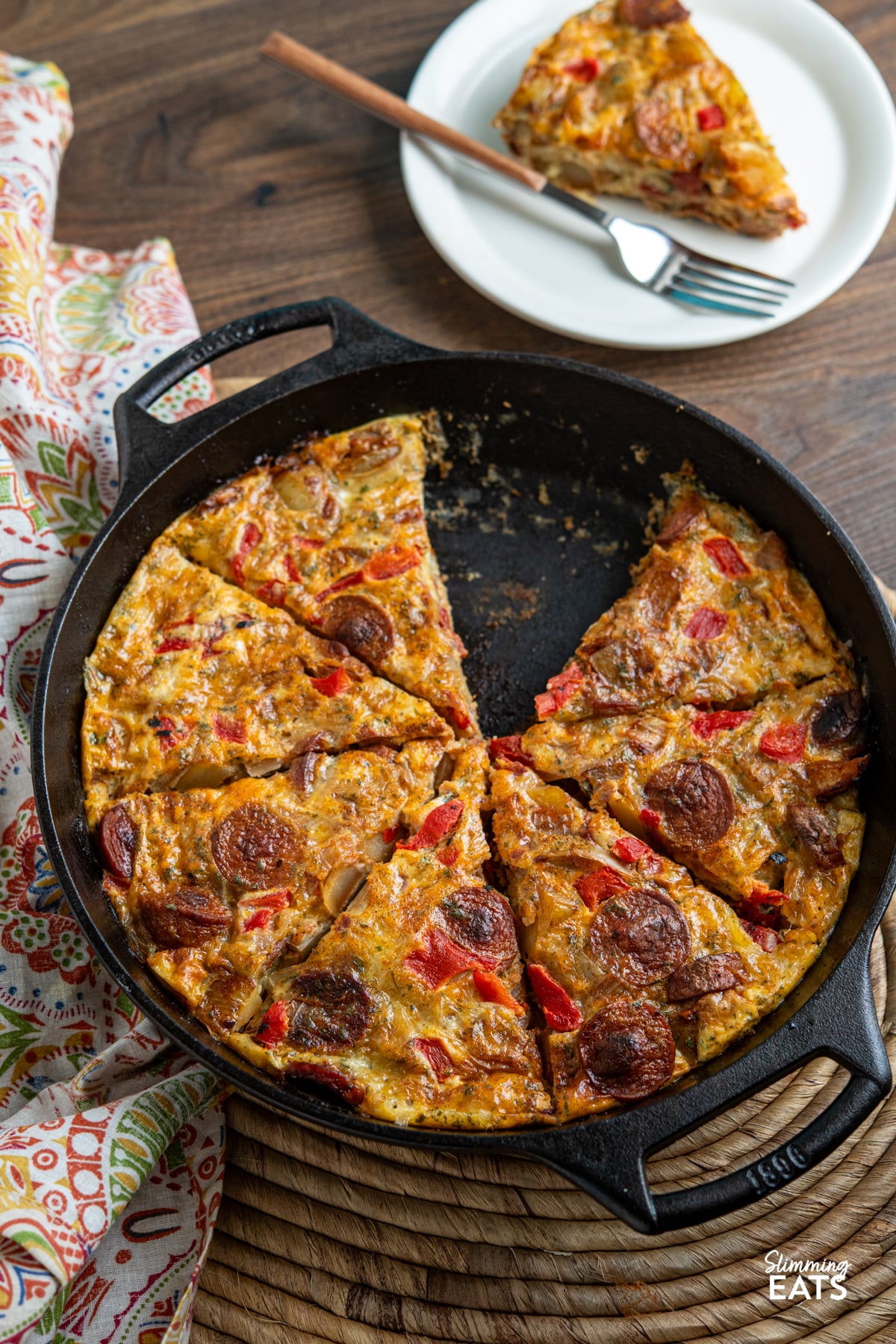Spicy smoked sausage, tender potato, roasted red pepper and cheddar come together in this easy frittata that is perfect for brunch, lunch or a simple dinner. sliced Spicy Smoked Sausage and Potato Frittata in a double handled blakc cast iron skillet with slice on a white plate with fork above