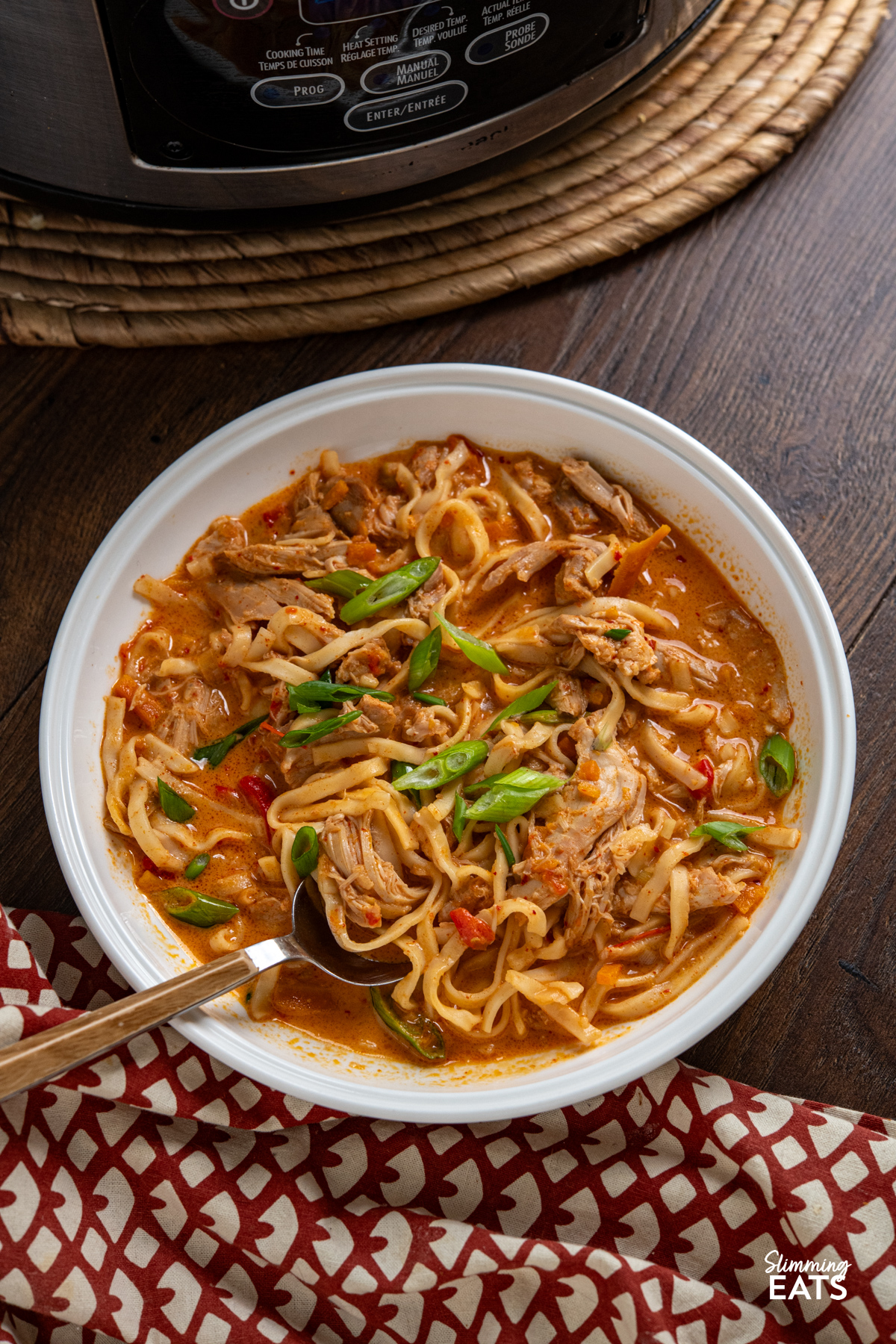 Slow Cooker Coconut Gochujang Chicken Noodles in a white bowl with a spoon placed in bowl, rusty brown and beige pattern placemat underneath 