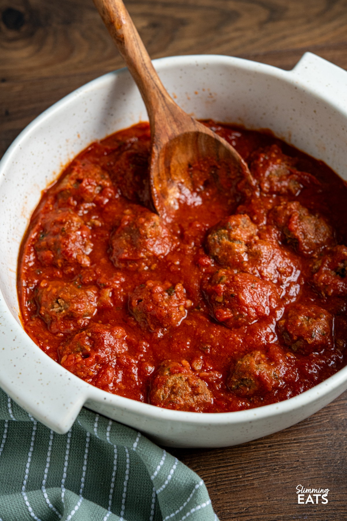 Slow Cooker Pesto Beef Meatballs are a comforting, flavour packed dinner with tender beef meatballs cooked in a rich tomato sauce. Slow Cooker Pesto Beef Meatballs in a white casserole dish with two handles, wooden spoon placed into sauce