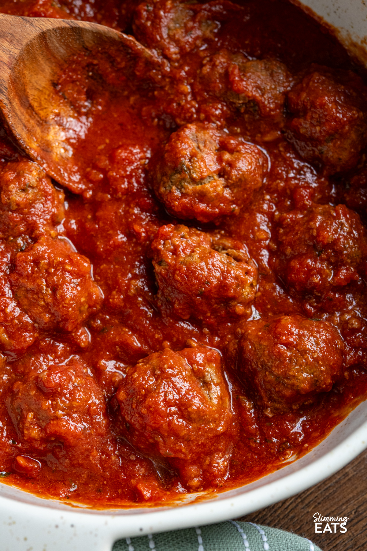 Slow Cooker Pesto Beef Meatballs are a comforting, flavour packed dinner with tender beef meatballs cooked in a rich tomato sauce. close up of Slow Cooker Pesto Beef Meatballs in a white casserole dish with two handles, wooden spoon placed into sauce